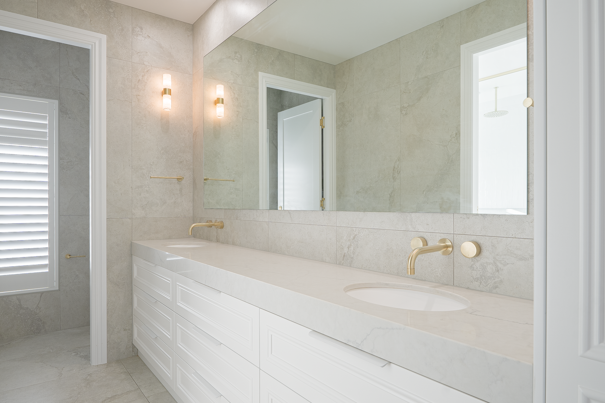 Modern bathroom with a large mirror, beige tiled walls, and a white vanity with a marble countertop. There are gold fixtures including a faucet, towel bar, and wall sconces. A window with white shutters lets in natural light, and part of a walk-in sh
