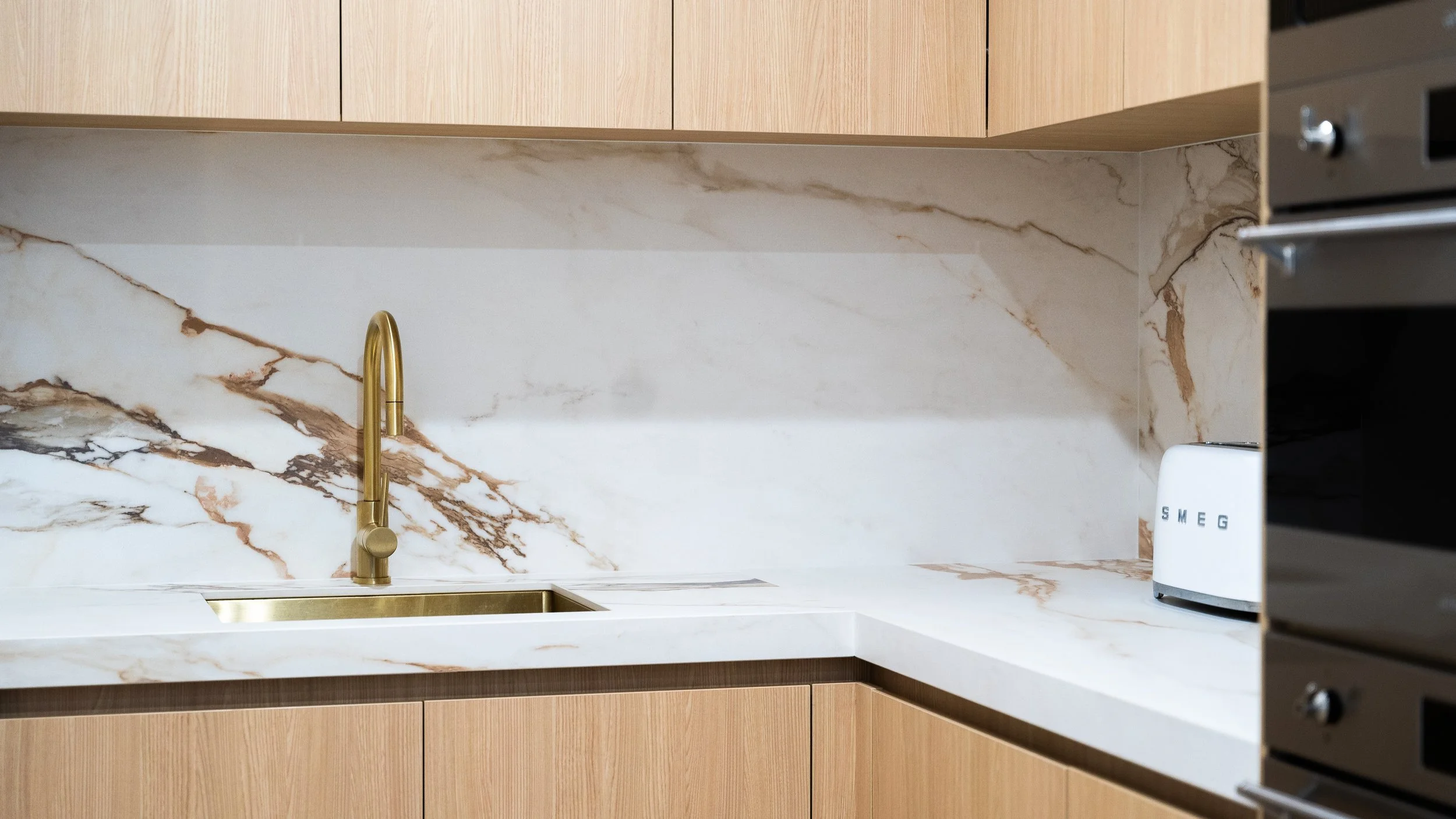 Kitchen with a marble backsplash, wood cabinets, a gold sink faucet, a white SMEG toaster, and a glimpse of a stainless steel oven.