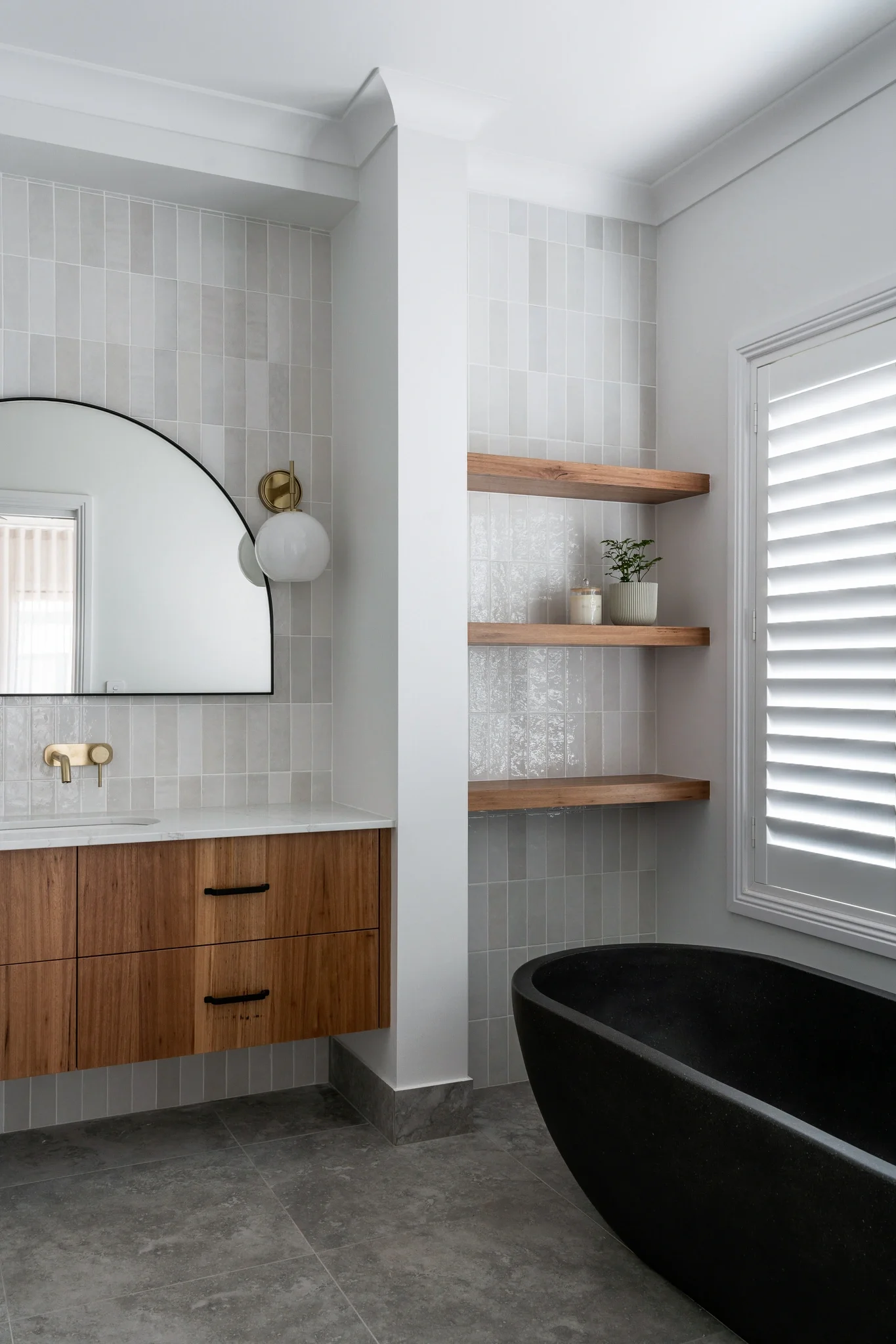 Modern bathroom with a black bathtub, wooden vanity, large mirror, white wall tiles, floating wooden shelves with a plant and candle, window with shutters, and gray tiled flooring.