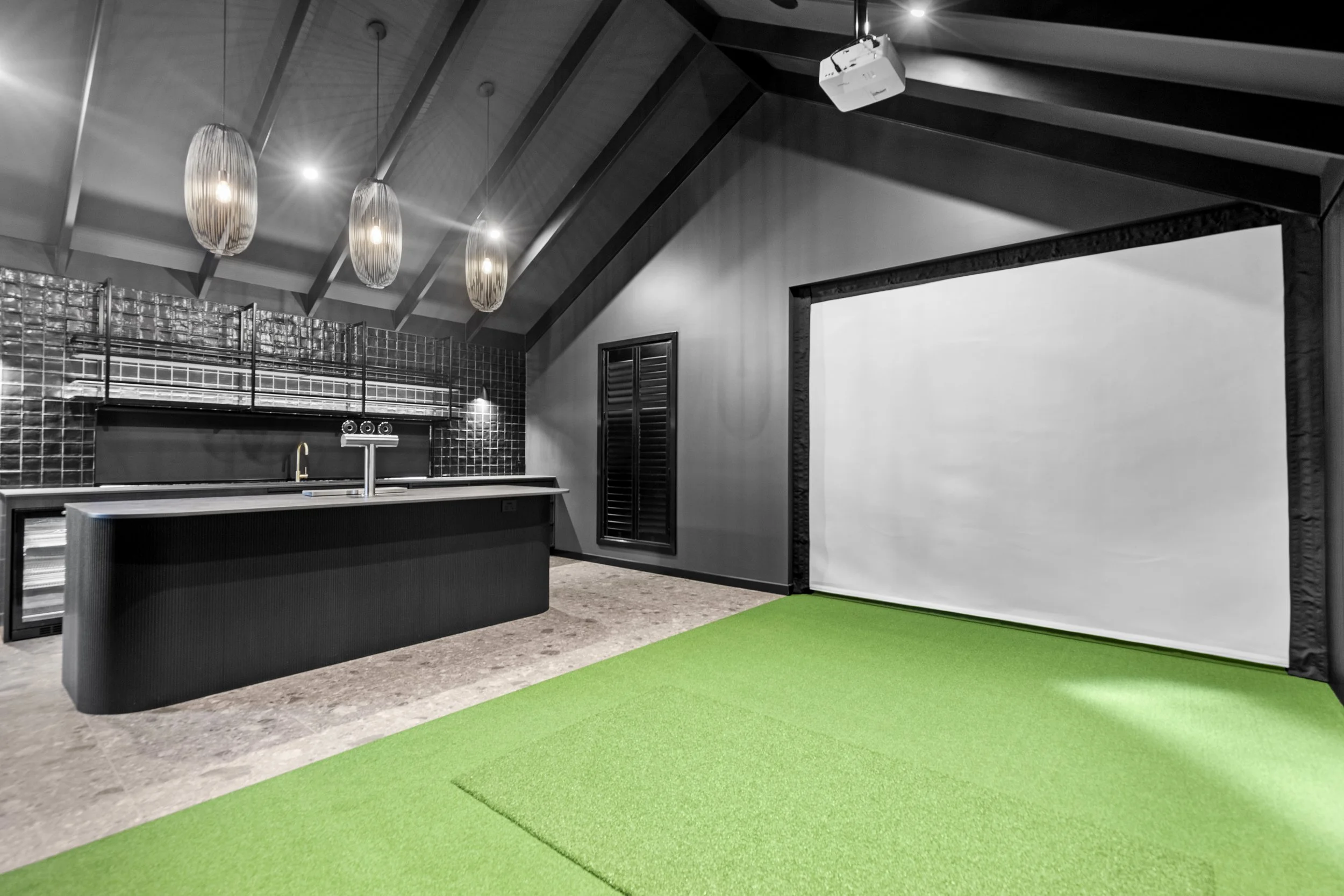 An indoor space with a green carpeted area, a black kitchen island, pendant lights, a black tiled wall, a window with black shutters, and a large white projection screen.