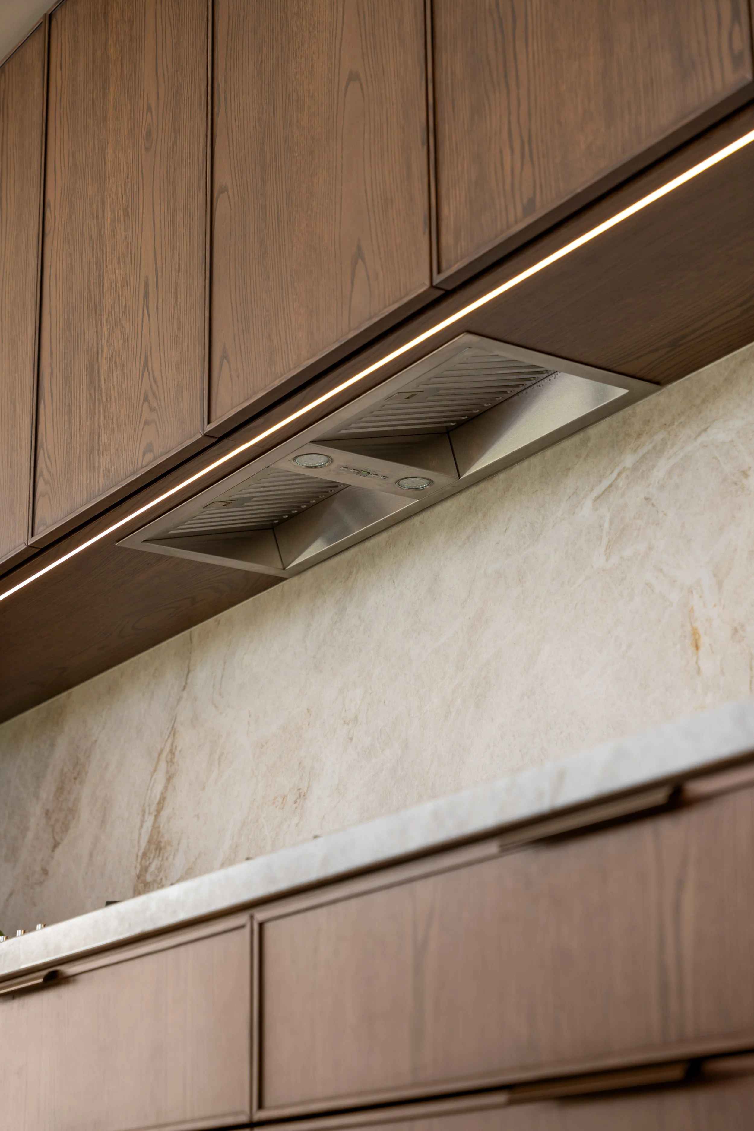 Close-up view of kitchen cabinets and a countertop with a built-in range hood underneath. The cabinets are made of wood with a natural finish, and there is a strip of LED lighting beneath the cabinets.