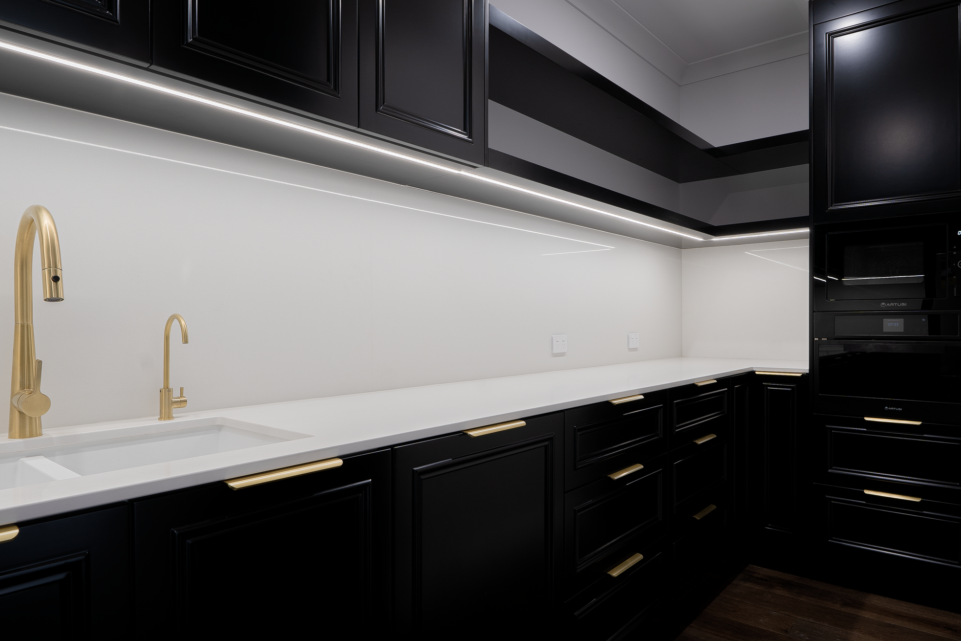 Modern kitchen with black cabinets, white countertops, gold fixtures, and built-in appliances, illuminated by under-cabinet lighting.