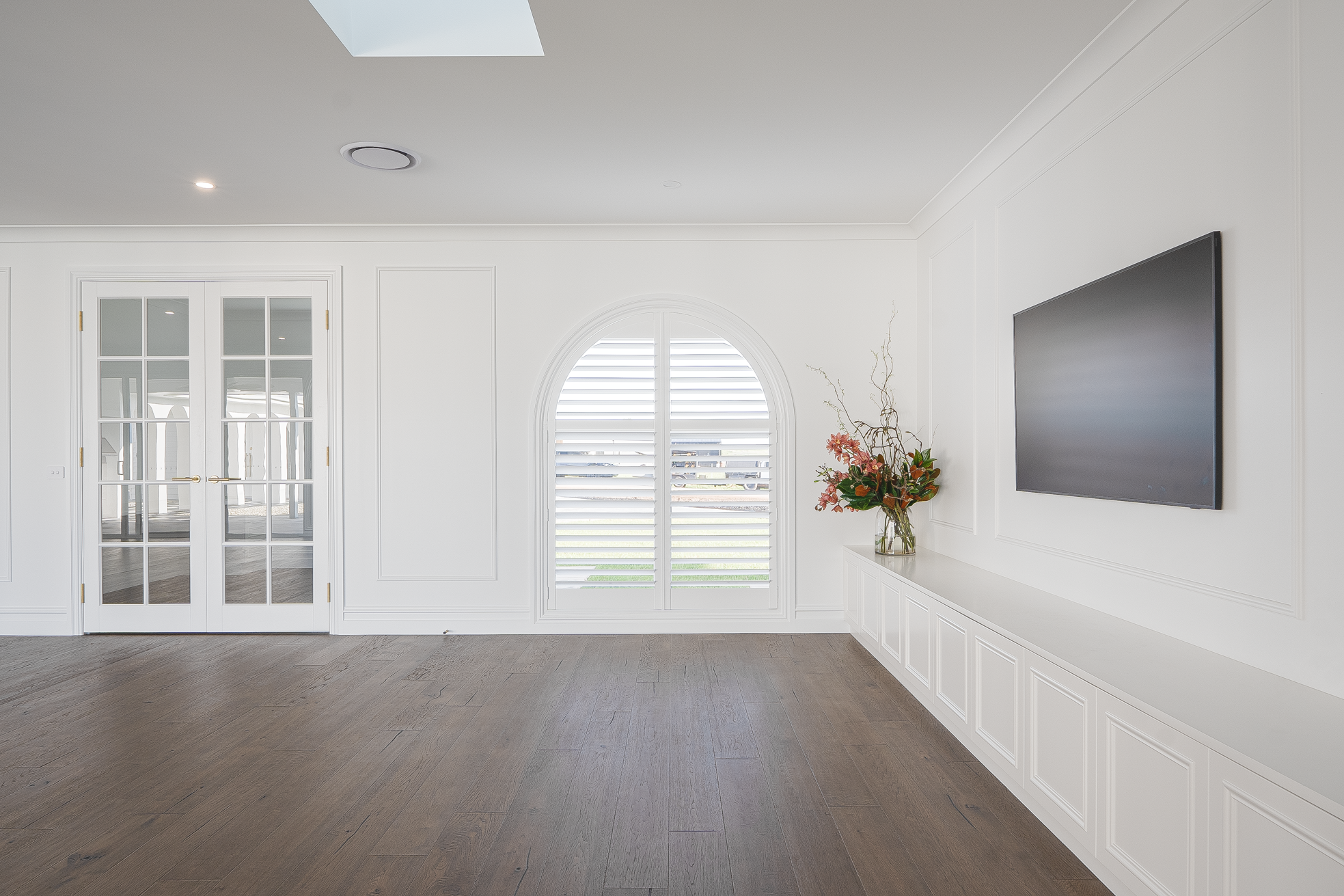 A bright living room with white walls, a large window with blinds, a mounted flat-screen TV, a glass vase with pink and orange flowers, and double glass doors leading outside.