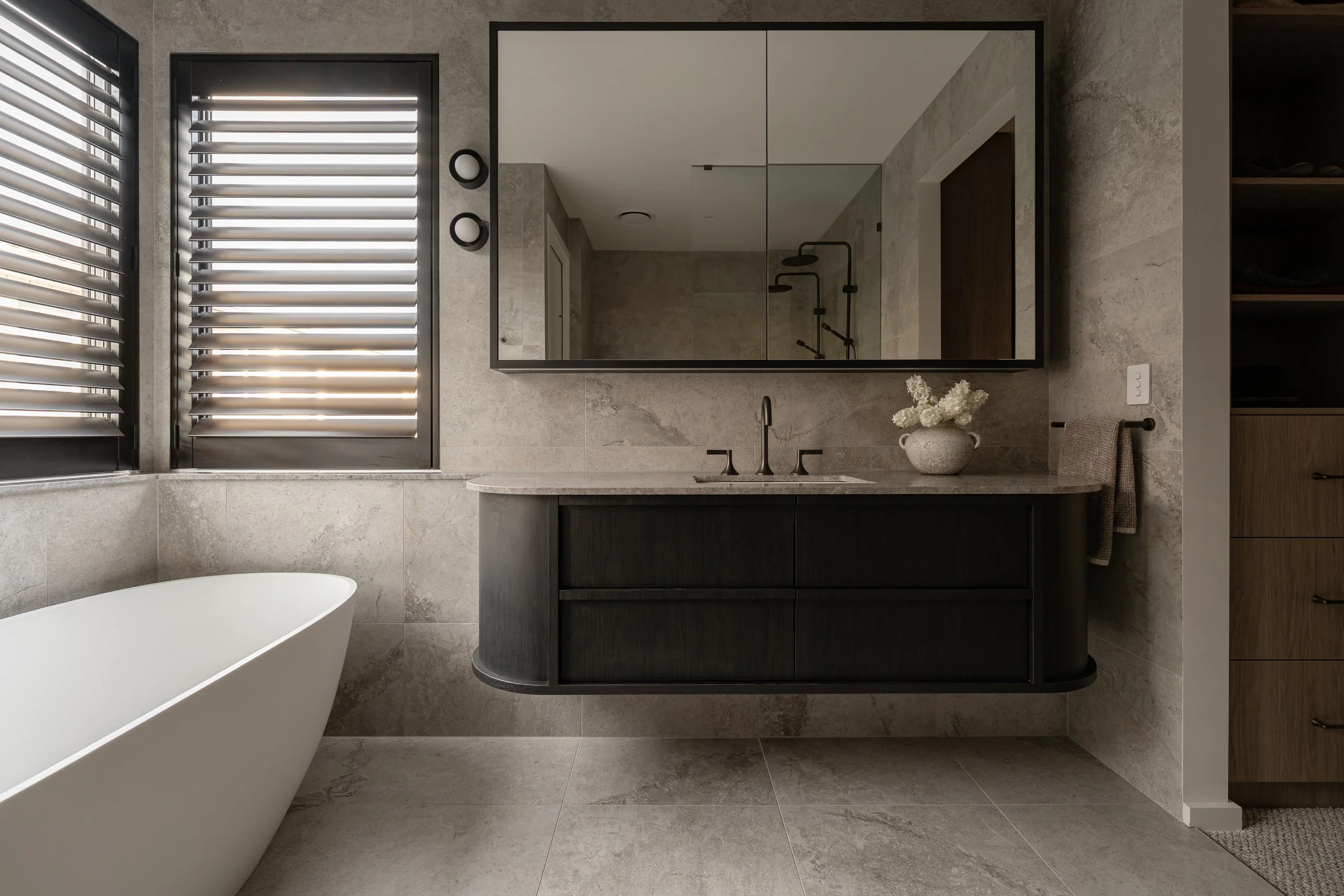 Modern bathroom with beige stone tiles, a large mirror, black fixtures, a white bathtub, and a vanity with a sink and decorative flowers.