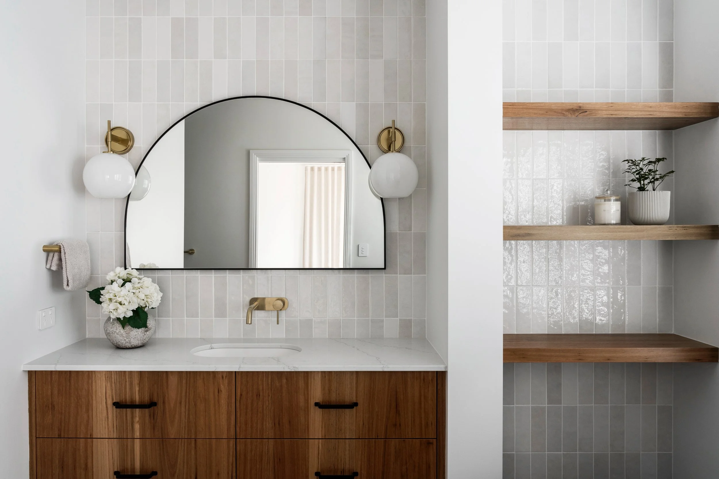 Close-up of a modern bathroom vanity with a white countertop, a gold faucet, and a wooden cabinet with black handles. There is a round mirror above the sink, flanked by two white globe wall sconces with gold bases. A vase of white flowers and a beige