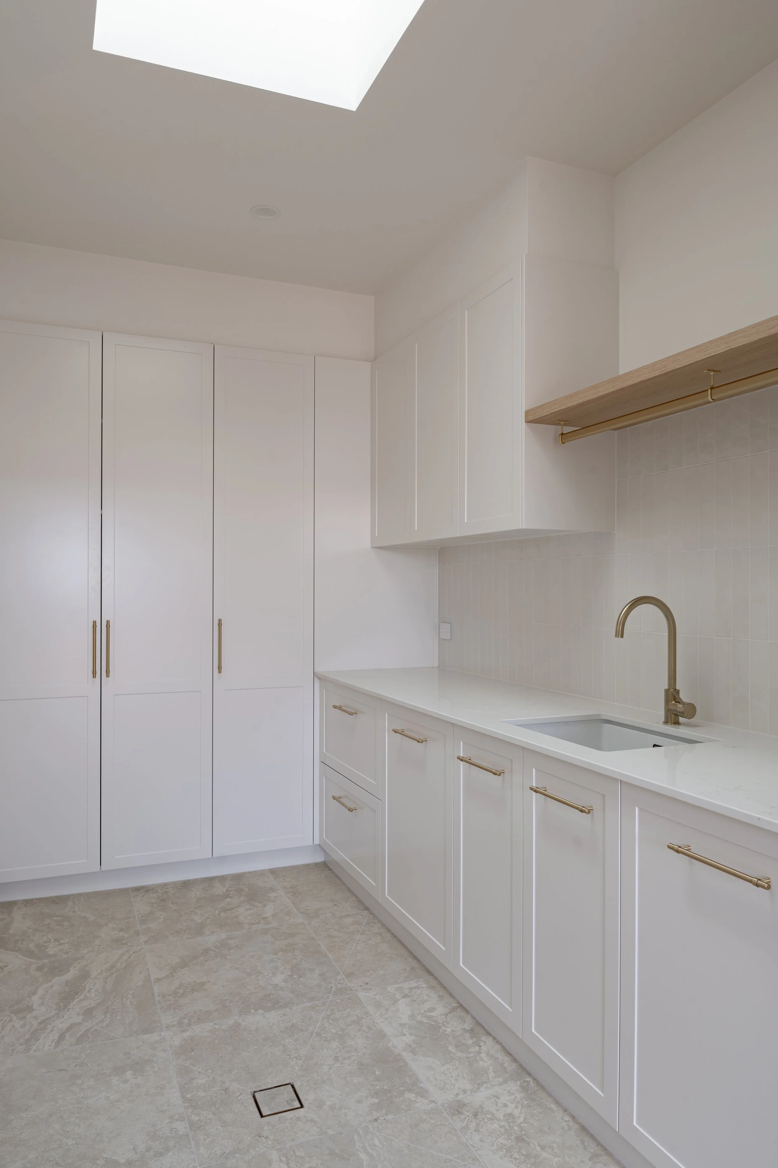 A clean, modern kitchen with white cabinets, gold handles, a white countertop, beige tiled flooring, a gold faucet, a sink, a wooden shelf with gold brackets, and a ceiling skylight.