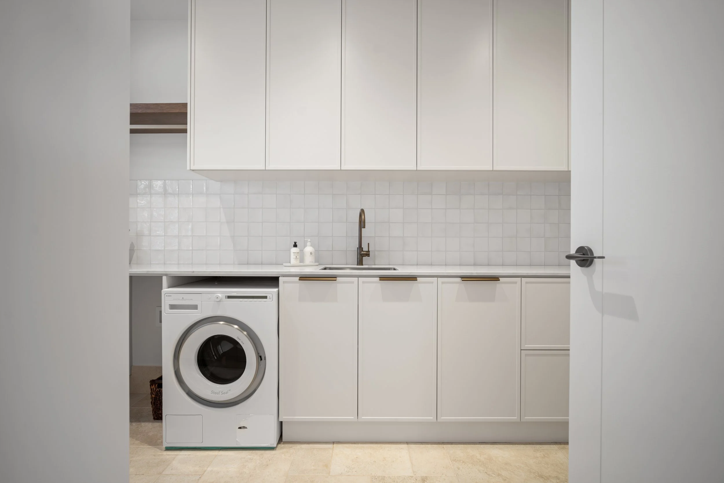 A laundry room with white cabinets above a white countertop, a sink with a brass faucet, and a tiled backsplash. There is a washing machine and two bottles of soap on the counter, seen through a partially open door.