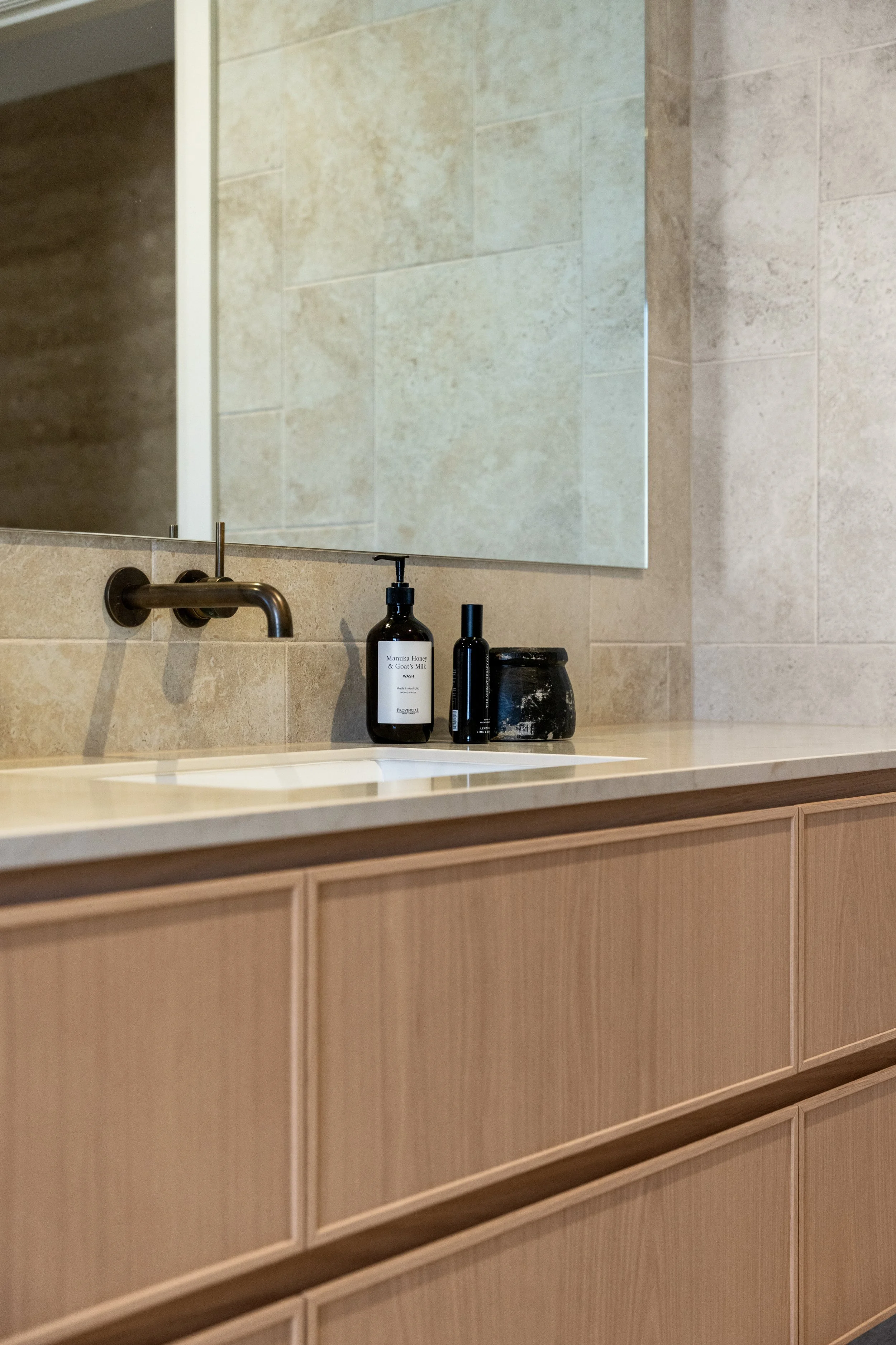 Bathroom vanity with a mirror, a wall-mounted faucet, and two bottles of skincare or soap products on the beige countertop.
