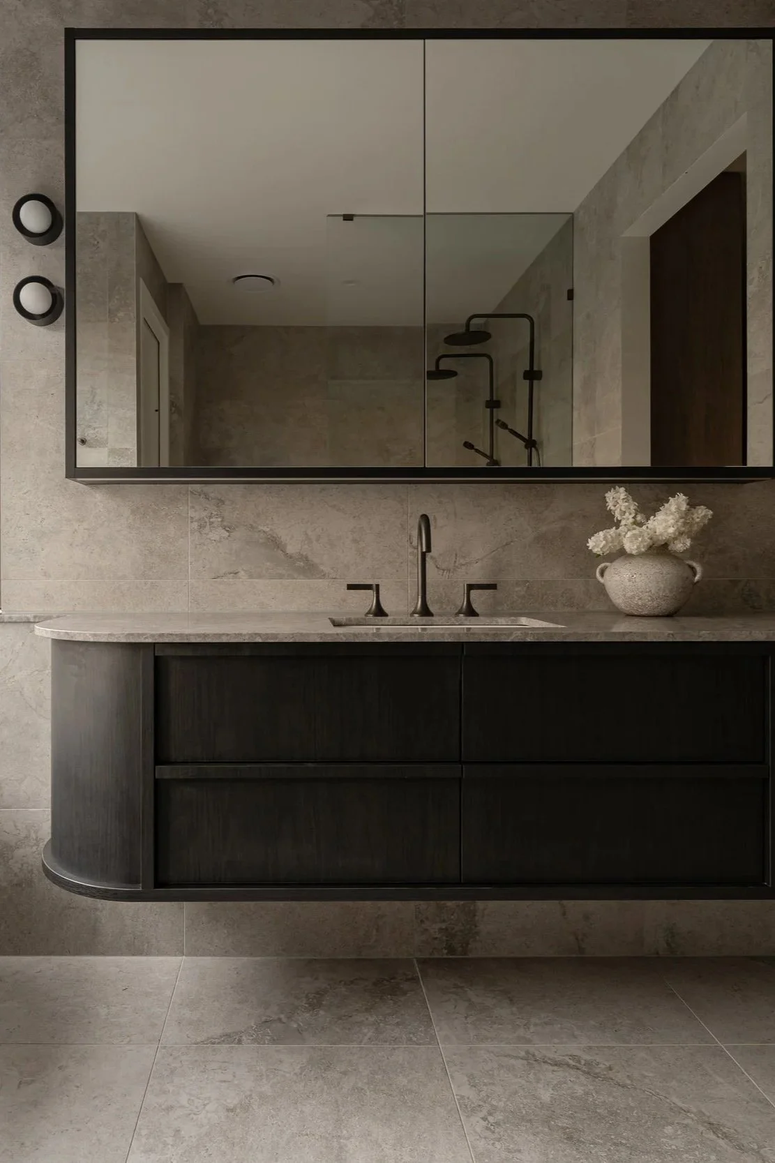 Modern bathroom with a large mirror, dark vanity with a marble countertop, a vase with white flowers, and a reflection of a shower with a rain showerhead in a walk-in shower area.