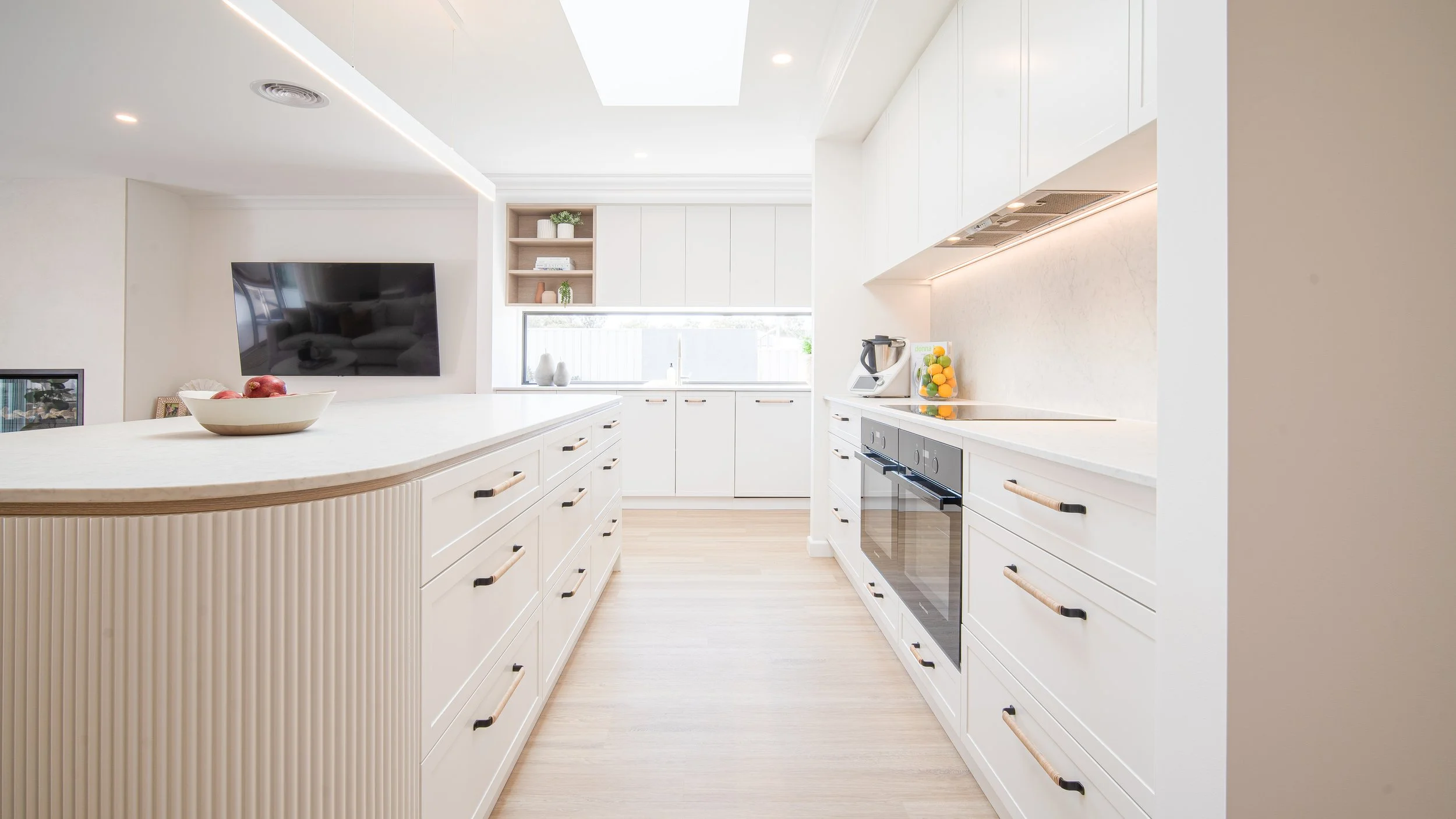 Modern, bright kitchen with white cabinets, countertops, and appliances, including an oven and blender, with a window and decorative items.