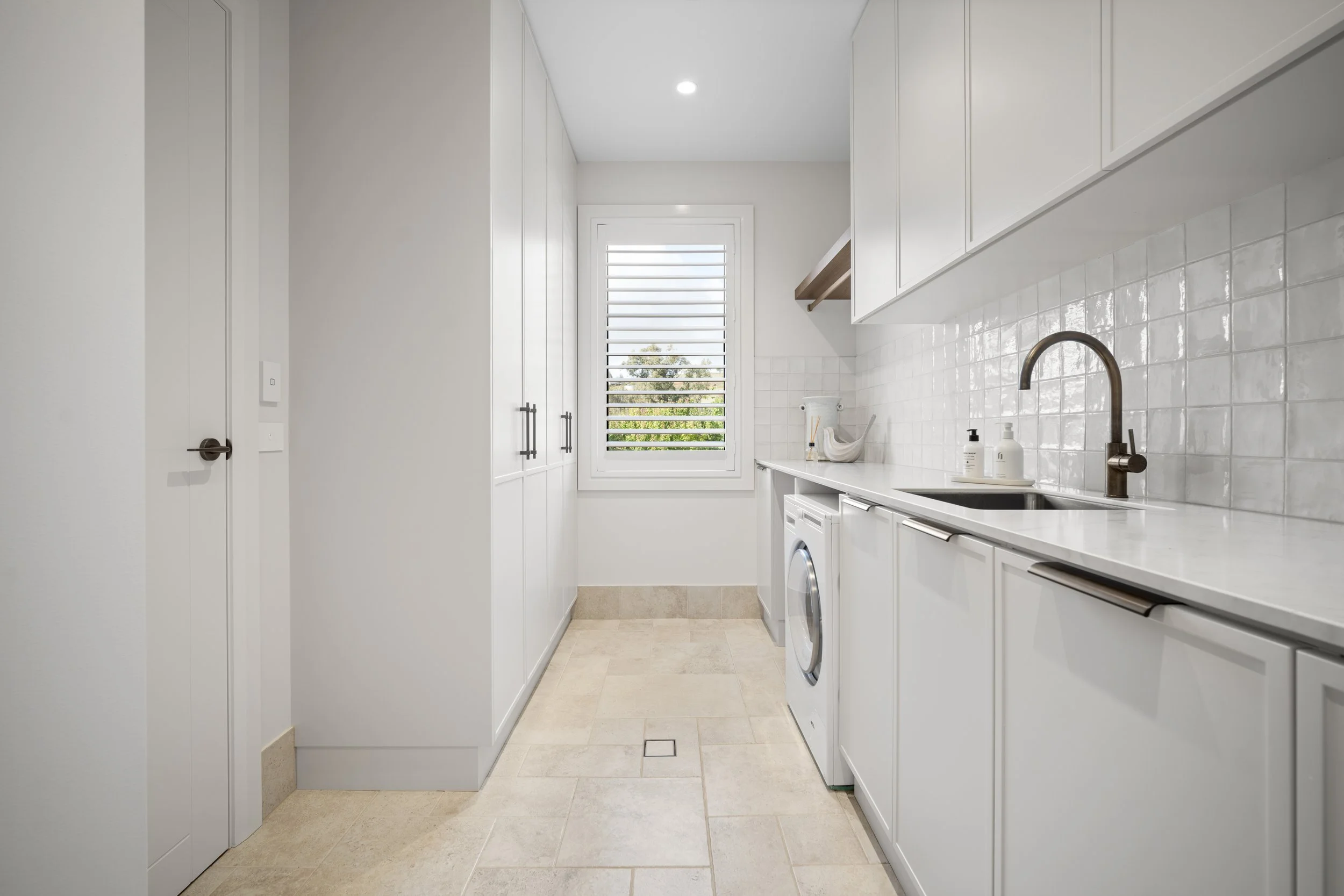 A modern, minimalist laundry room with white cabinetry, a window with shutters, a washing machine, and a small shelf with decor.