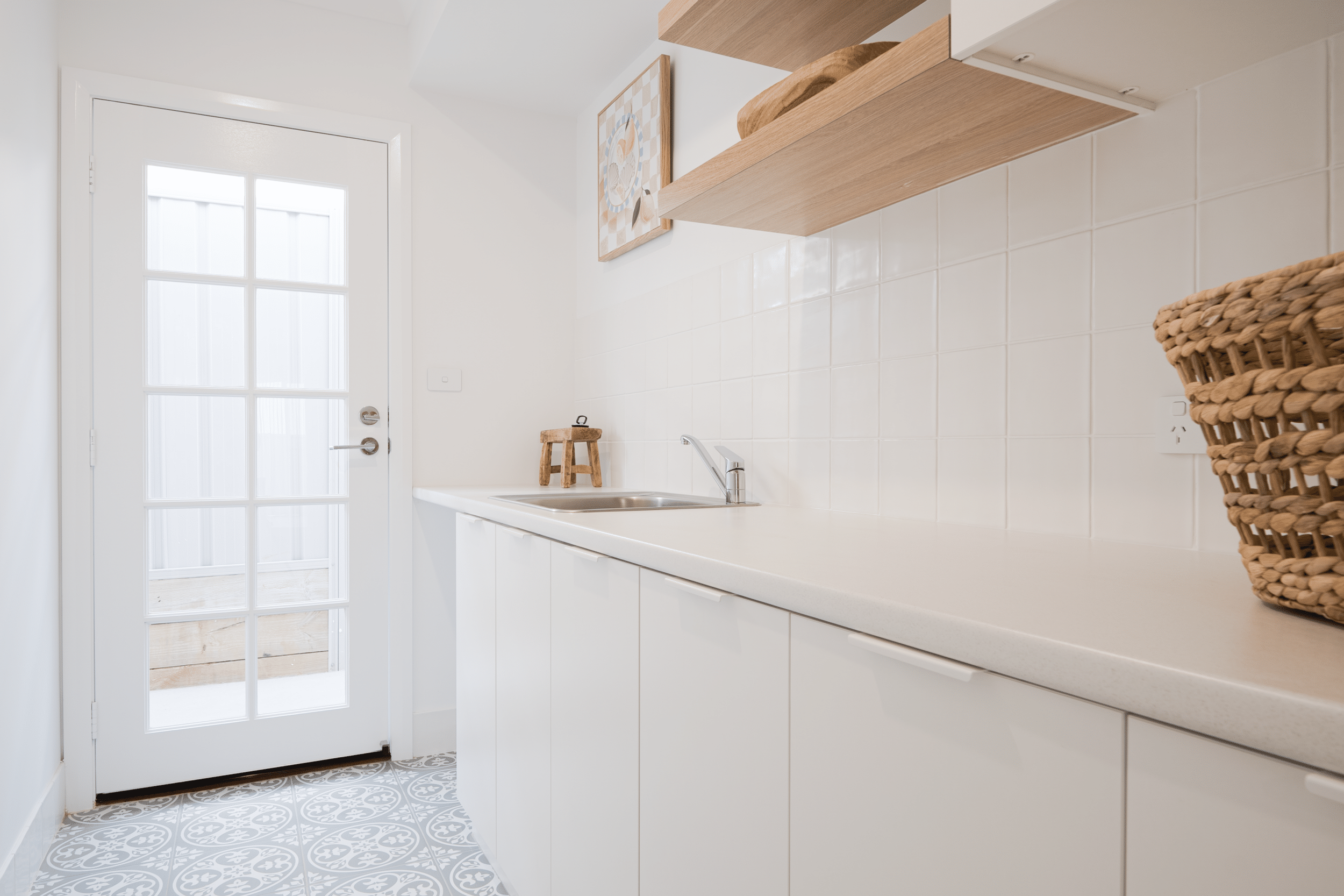 A bright kitchen with white cabinets, a stainless steel sink, and a wooden shelf. There is a wicker basket on the counter, and a door with glass panes leads outside. The floor has patterned tiles, and a small wooden stool is near the sink. Two framed