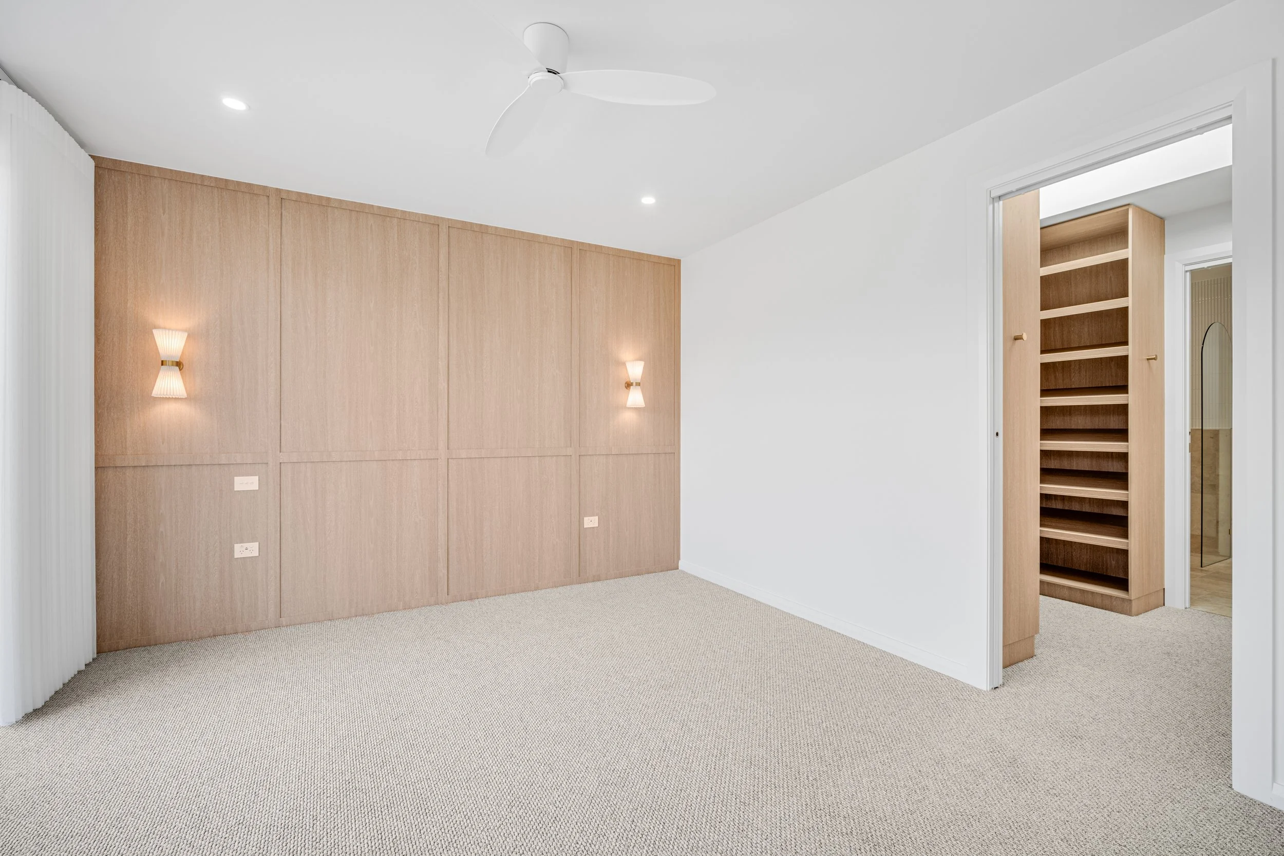Empty room with beige carpeting, white walls, wooden paneling, a built-in wooden shelving unit, ceiling fan, and wall-mounted lights.