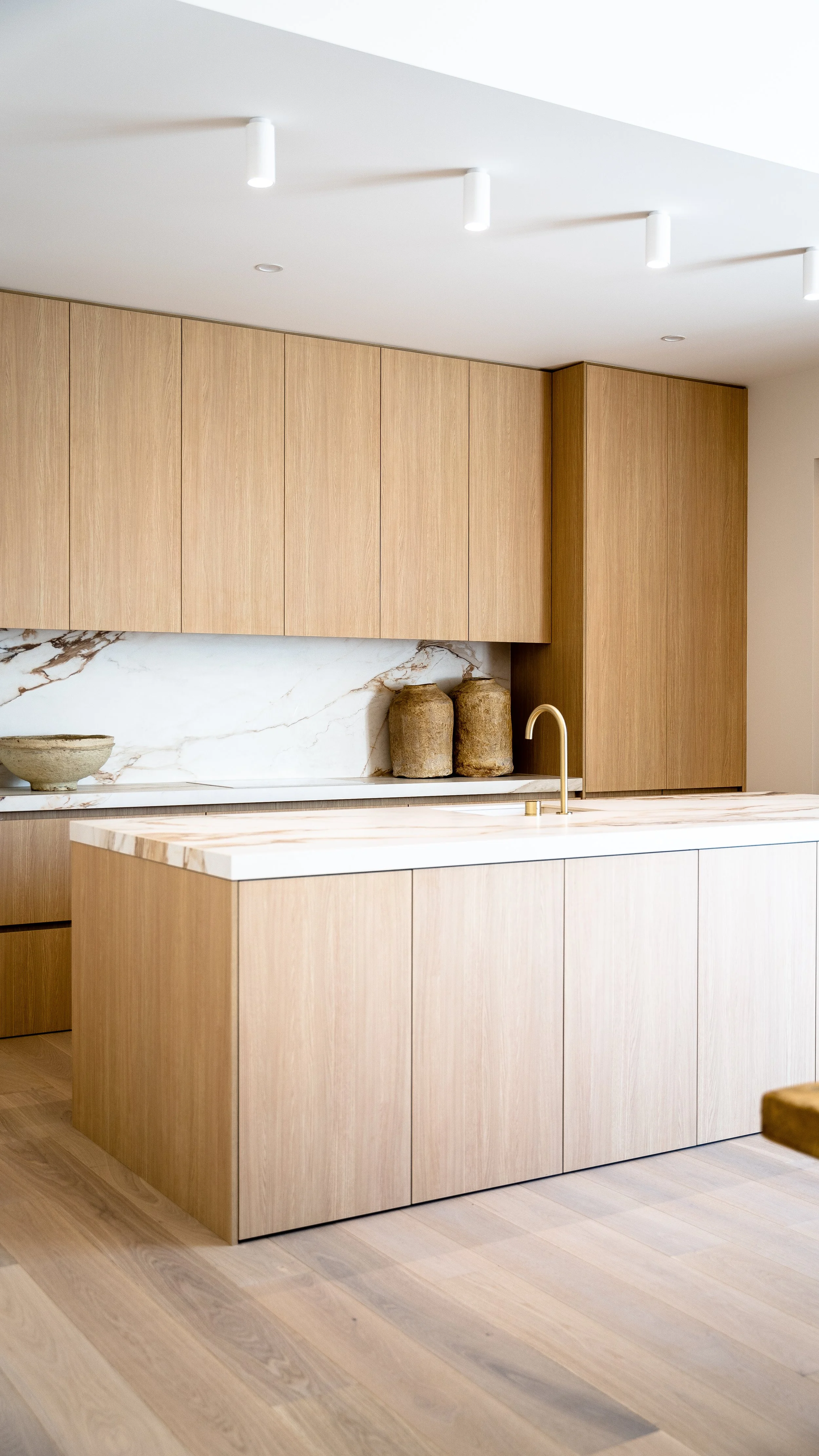 Modern kitchen with light wood cabinets, marble backsplash, and island with a gold faucet.
