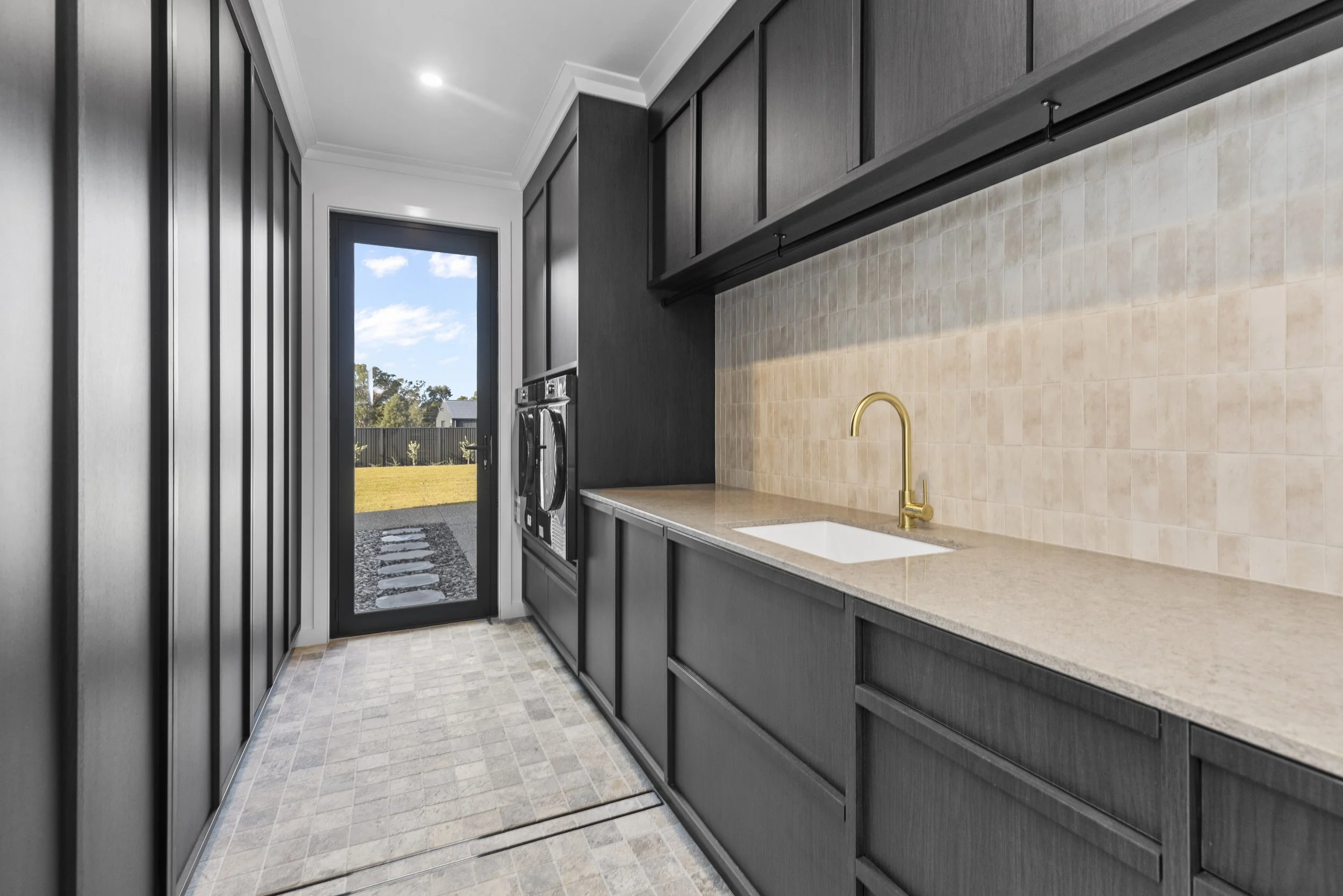 Modern kitchen with dark cabinets, beige countertop, brass faucet, beige tiled backsplash, and a glass door leading outside to a backyard with a fence and trees.