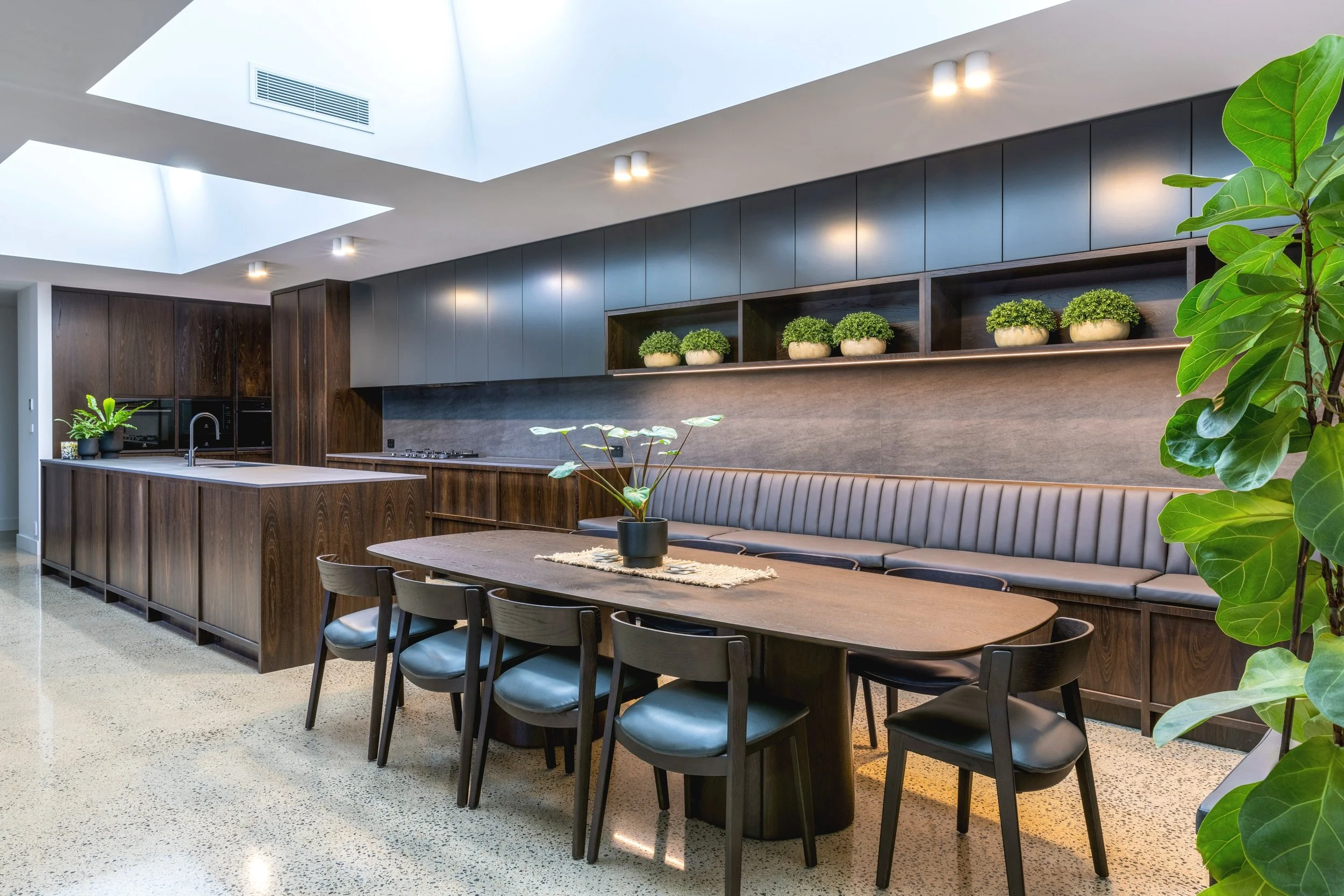 Modern kitchen and dining area with skylights, dark wood cabinets, a long dining table with black chairs, and green potted plants.