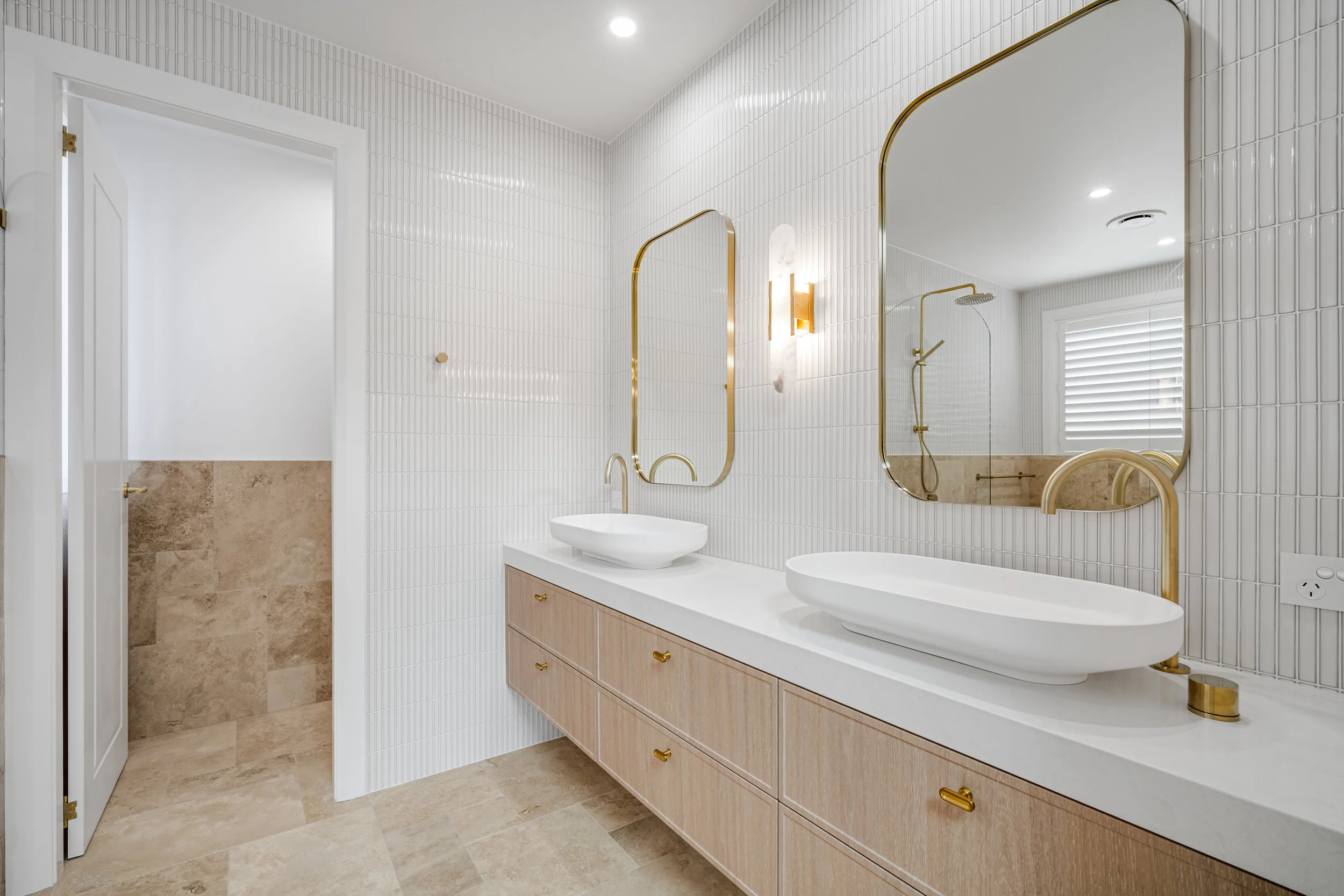 Modern bathroom with white tiled walls, a double sink vanity with a light wood cabinet and gold hardware, and a large mirror. There is a walk-in shower with beige tiles and a rainfall showerhead. A window with white blinds is reflected in the mirror,