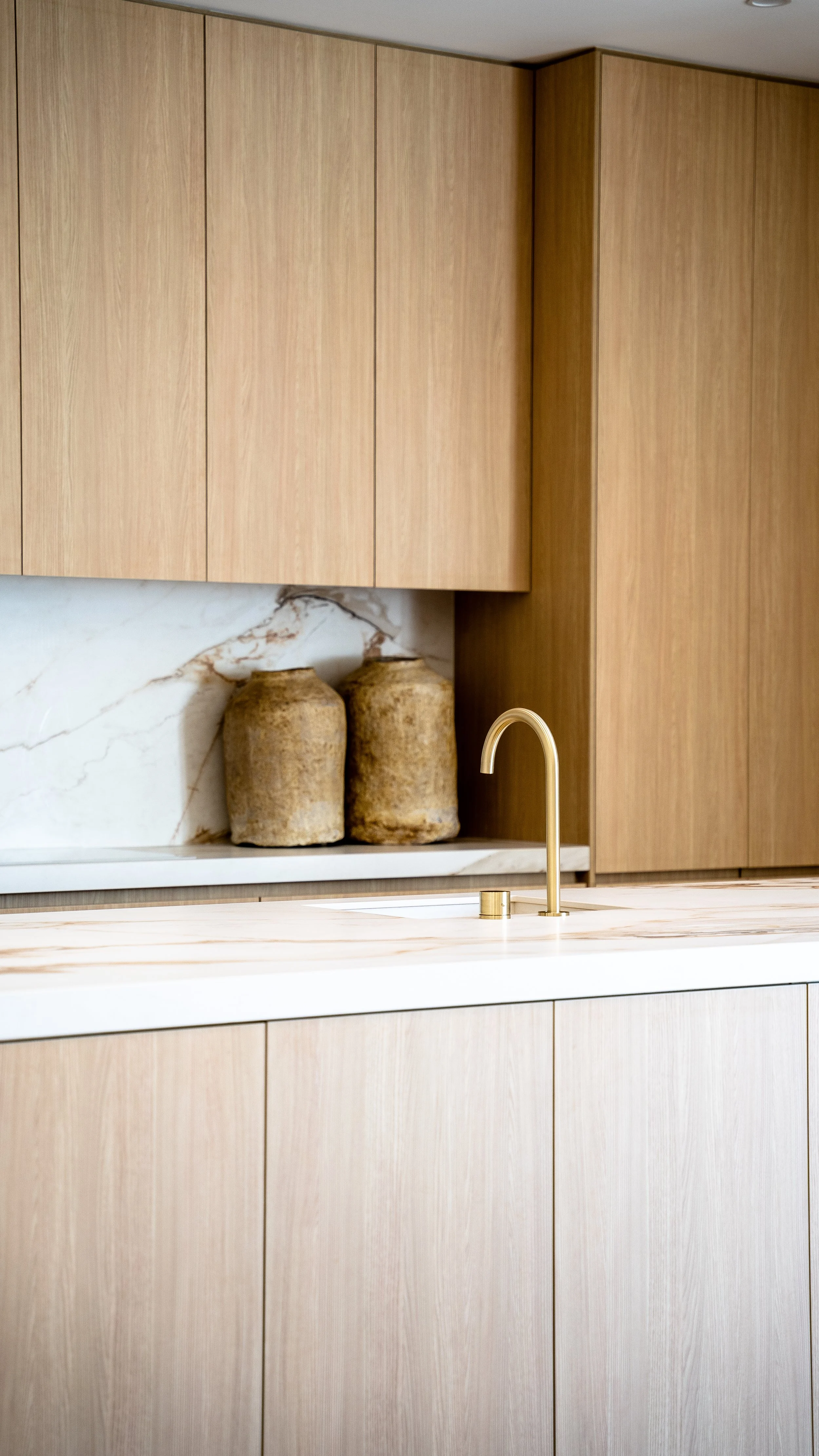 Modern kitchen with light wood cabinets, a white marble backsplash, a gold gooseneck faucet, and two decorative vases.