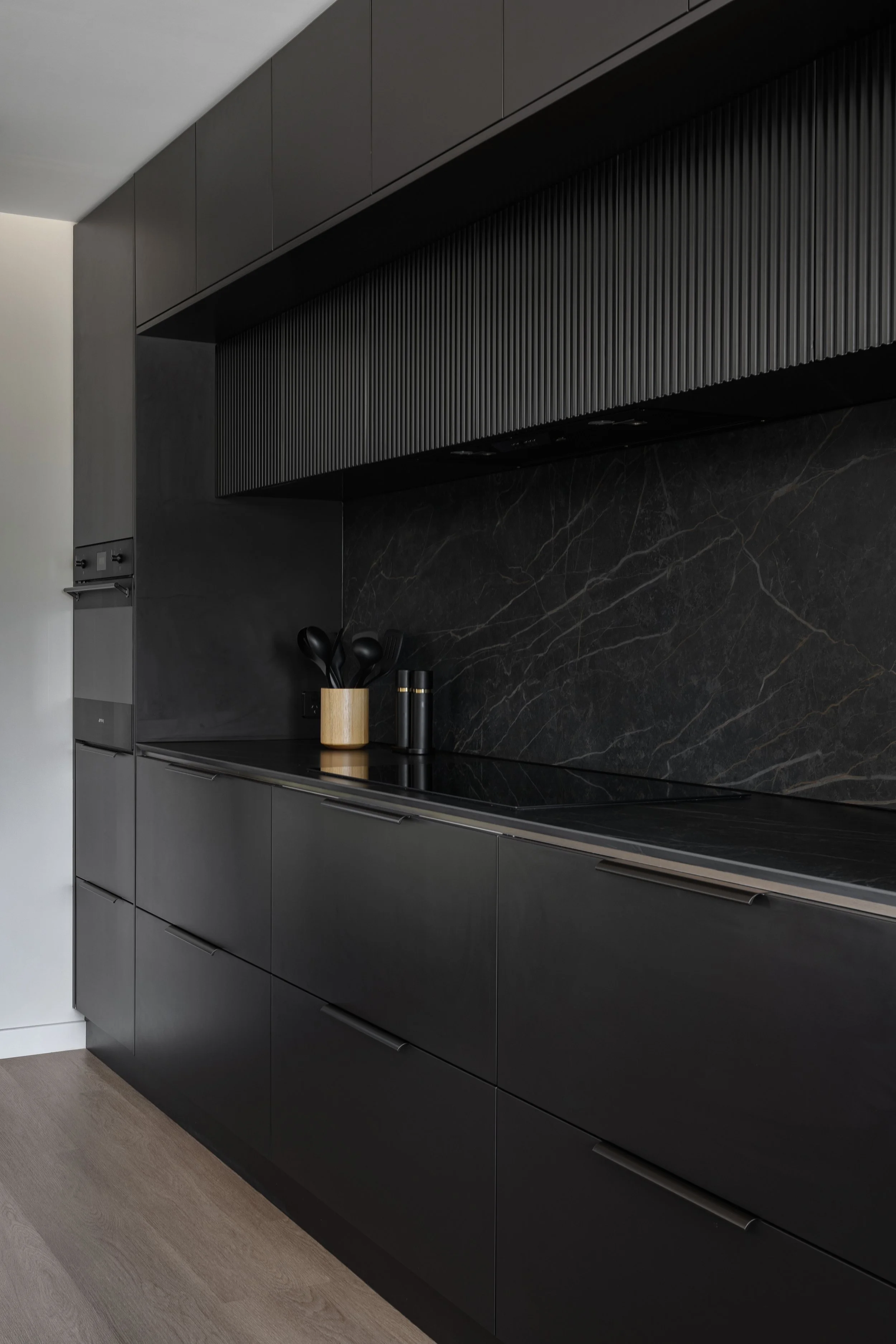 Modern kitchen with black cabinetry, black marble backsplash, matte black cabinet handles, and wooden utensil holder on black countertops.