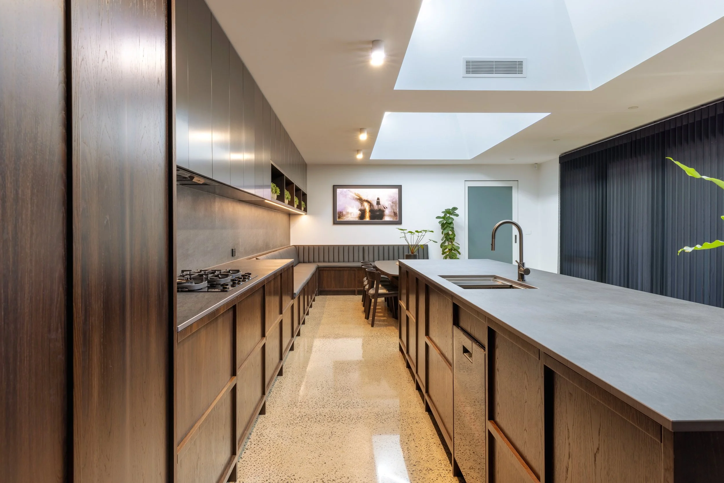 Modern kitchen with wooden cabinetry, an island with a sink, a seating area, framed art on the wall, and skylights in the ceiling.