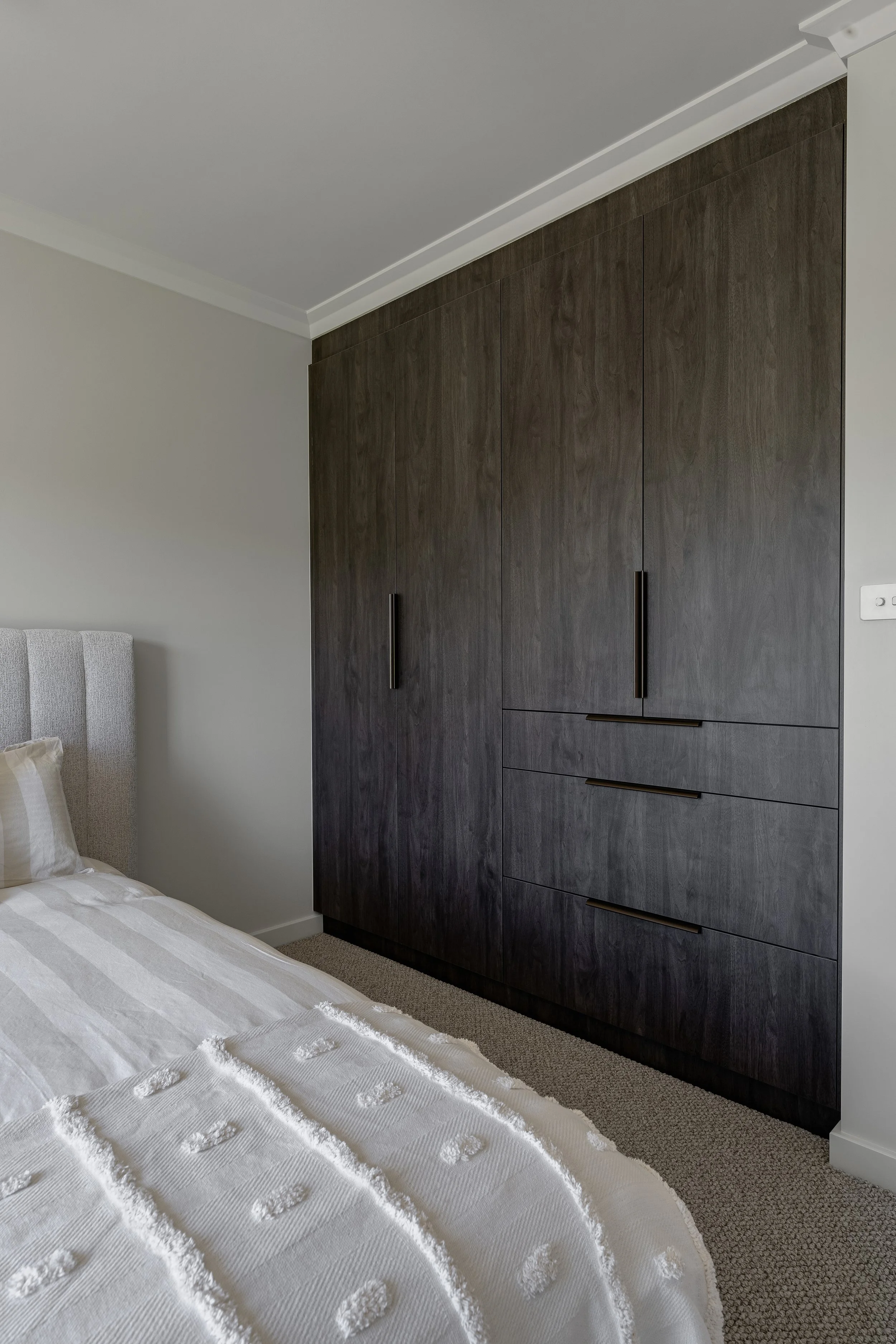 A bedroom with a white bedspread and a padded headboard. A large brown wooden wardrobe with vertical handles is next to the bed. The walls are painted light gray, and there is a white ceiling with crown molding.
