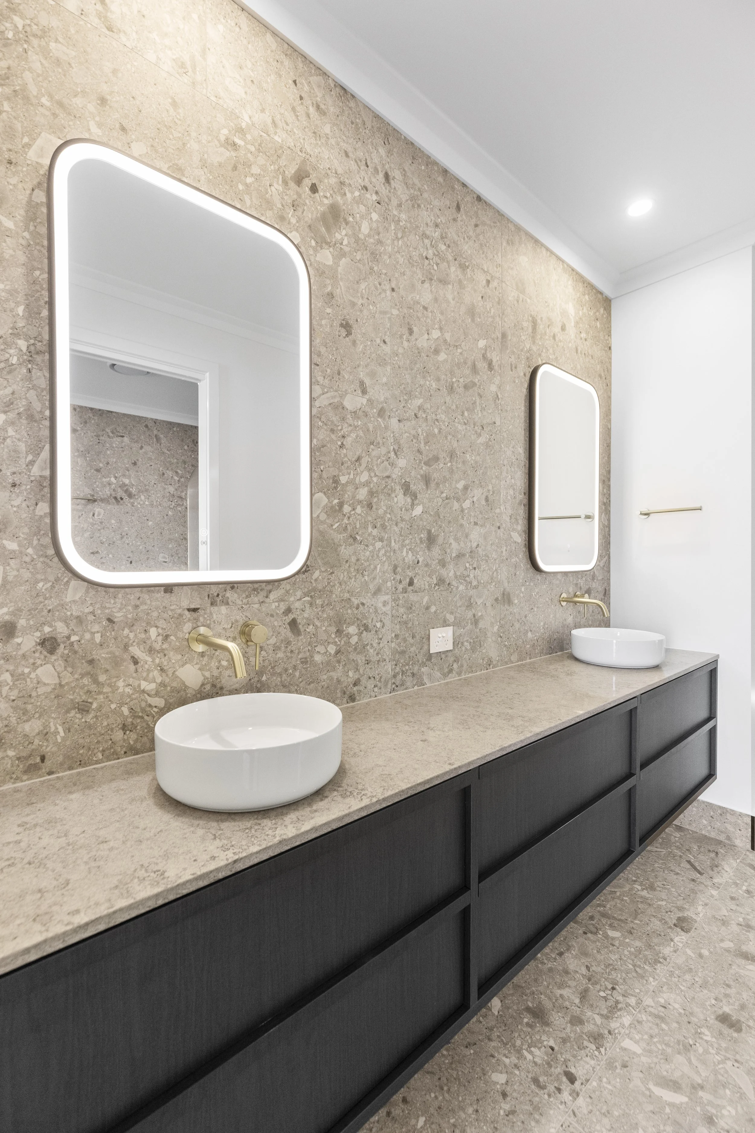 Modern bathroom vanity with two white vessel sinks on a beige stone countertop, gold fixtures, black cabinets, two rectangular mirrors with LED lighting, and beige tiled wall and floor.