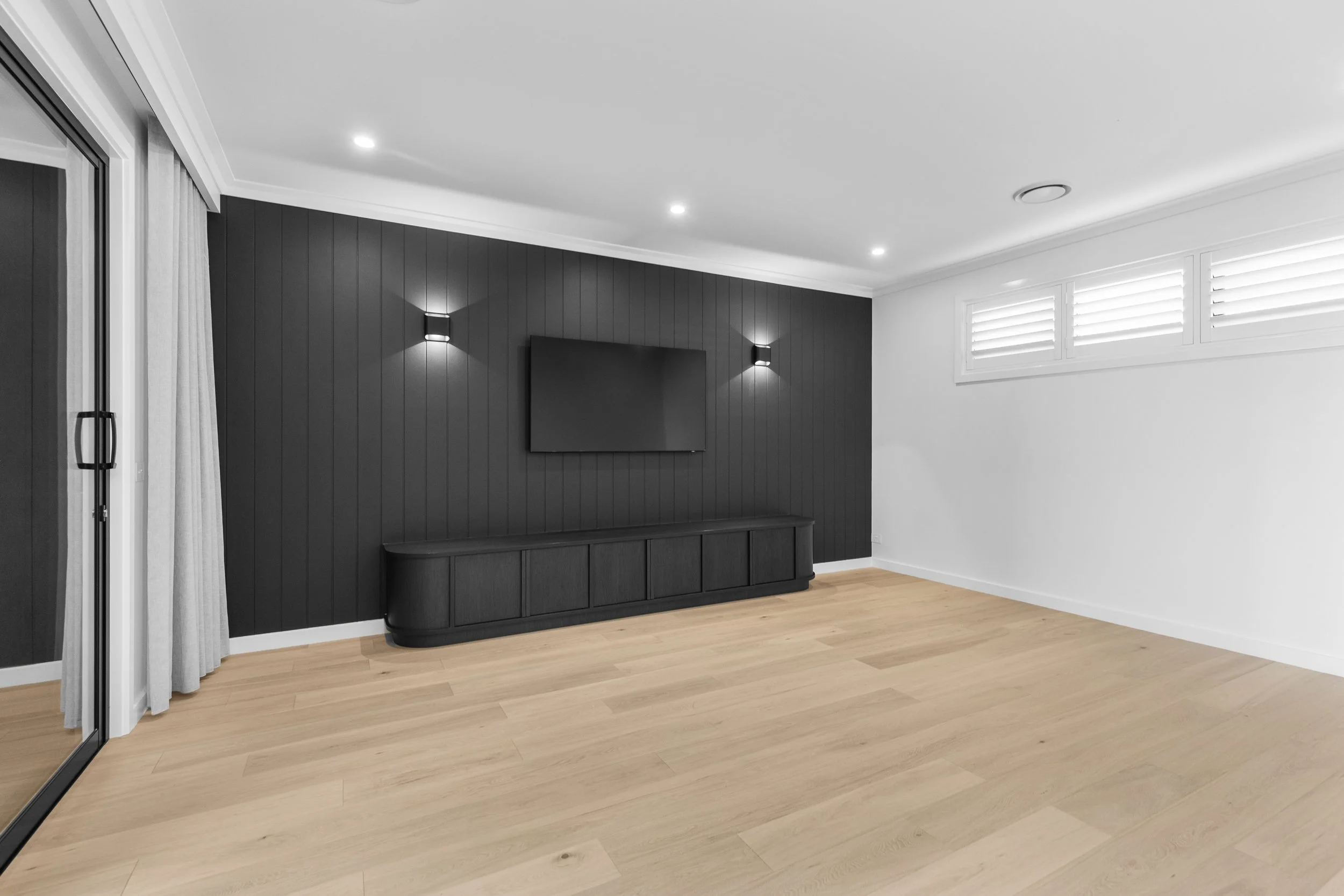Interior of a modern living room with wood flooring, white ceiling, black accent wall with mounted flat-screen TV, floor-length light gray curtains, and a window with white shutters.