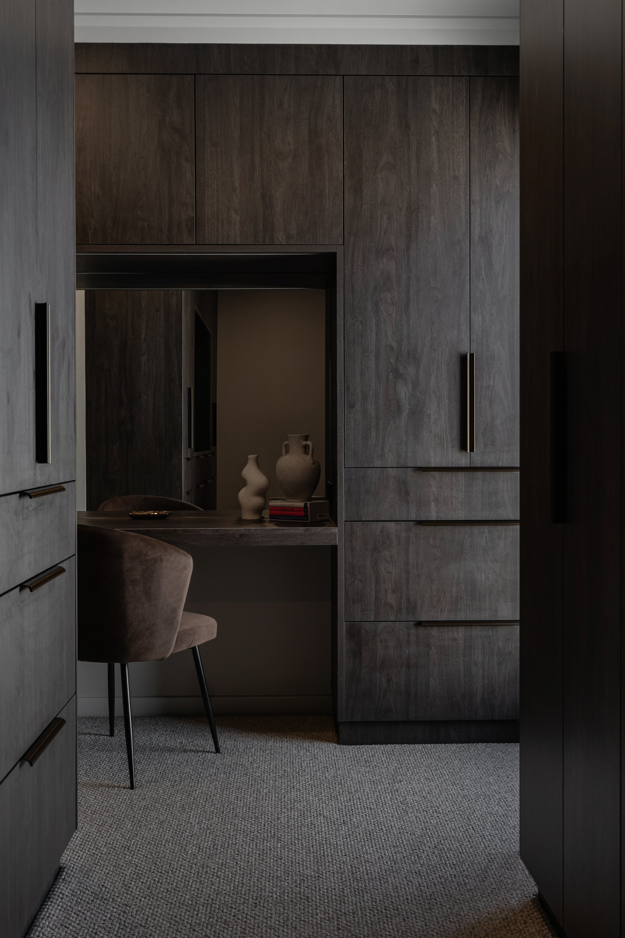 A glimpse into a modern, dark wood-paneled office or dressing area with a built-in desk and shelves. A beige upholstered chair is positioned in front of the desk, which has a few decorative vases and a stack of books. The area is carpeted with a text