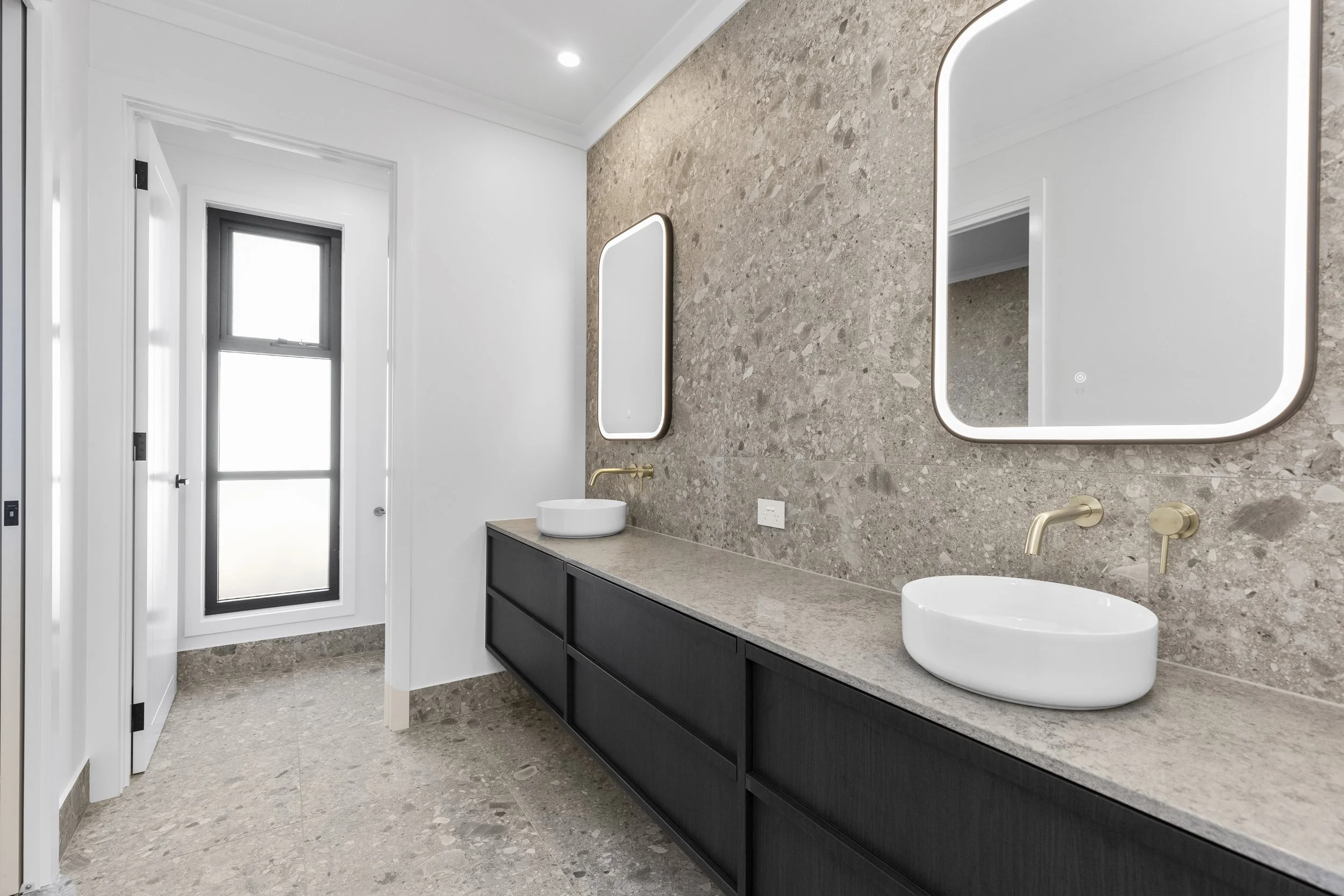 Modern bathroom with a double vanity, unique circular vessel sinks, large backlit mirrors, and beige stone wall and floor tiles.