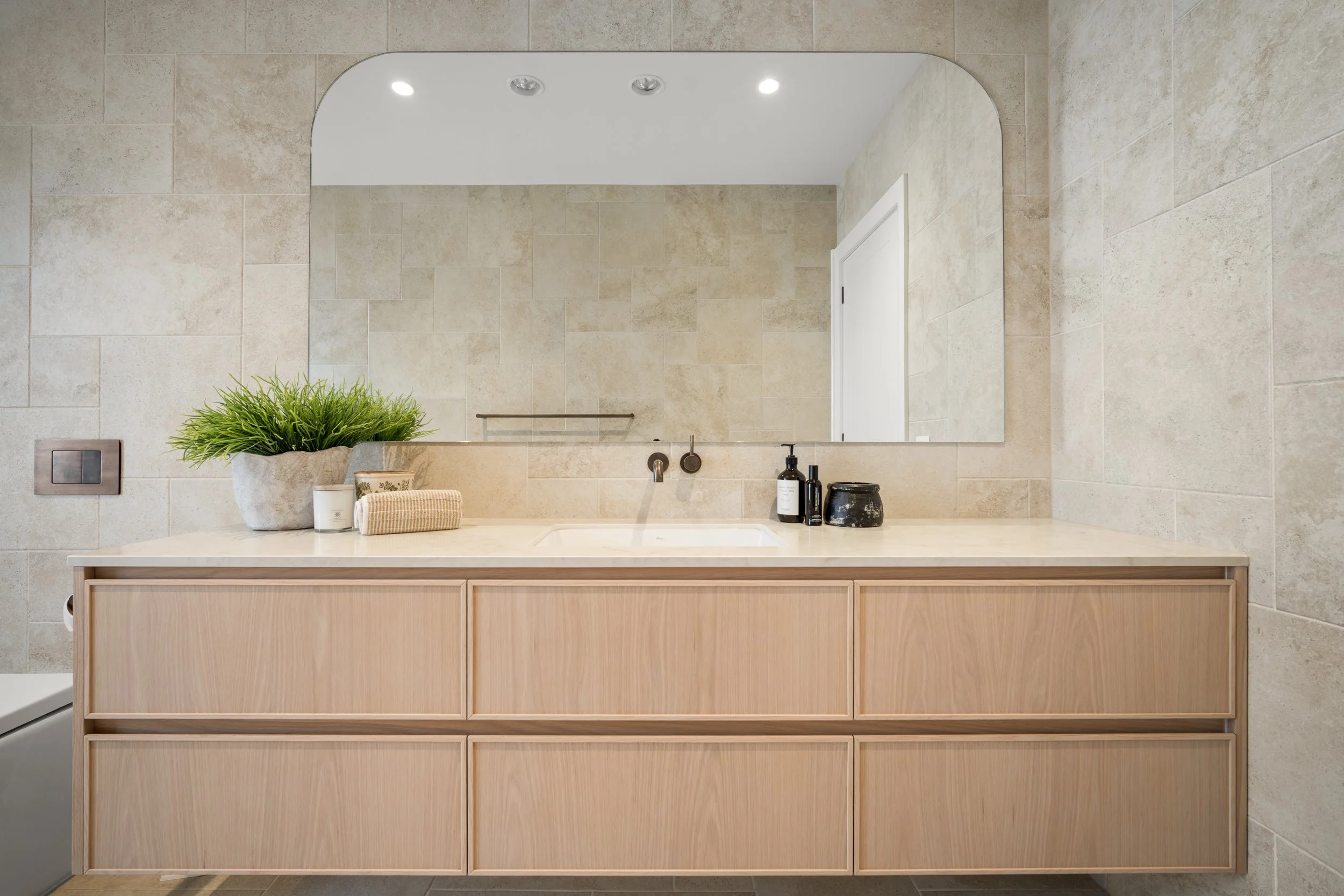 Bathroom vanity with a large mirror, beige tiled wall, potted green plant, soap dispenser, hairbrush, and black jar on a wooden cabinet.