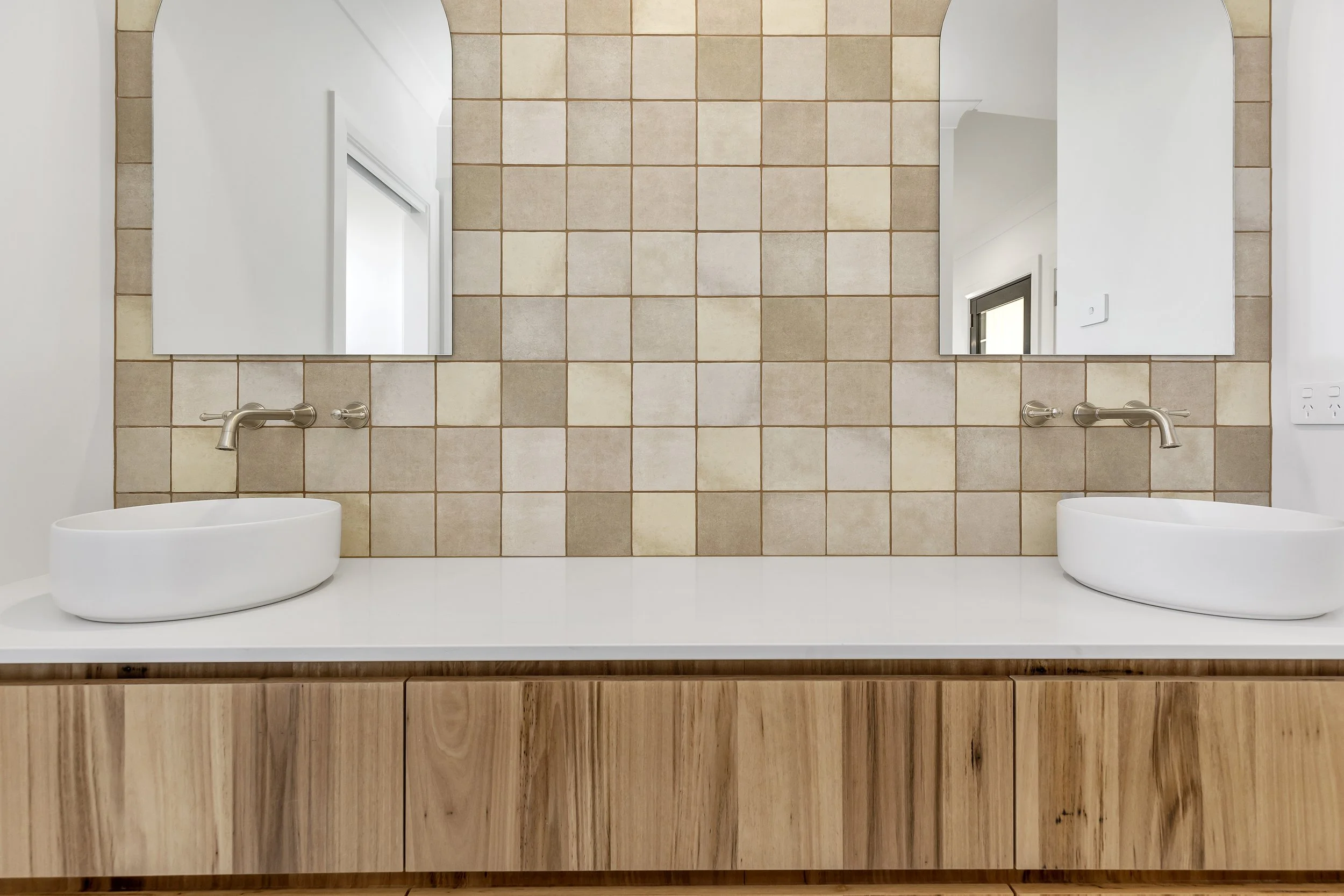 A bathroom vanity with a white countertop, two vessel sinks, and two mirrors above them. The wall behind the mirrors is covered with beige tiles, and the vanity cabinet is made of light wood.
