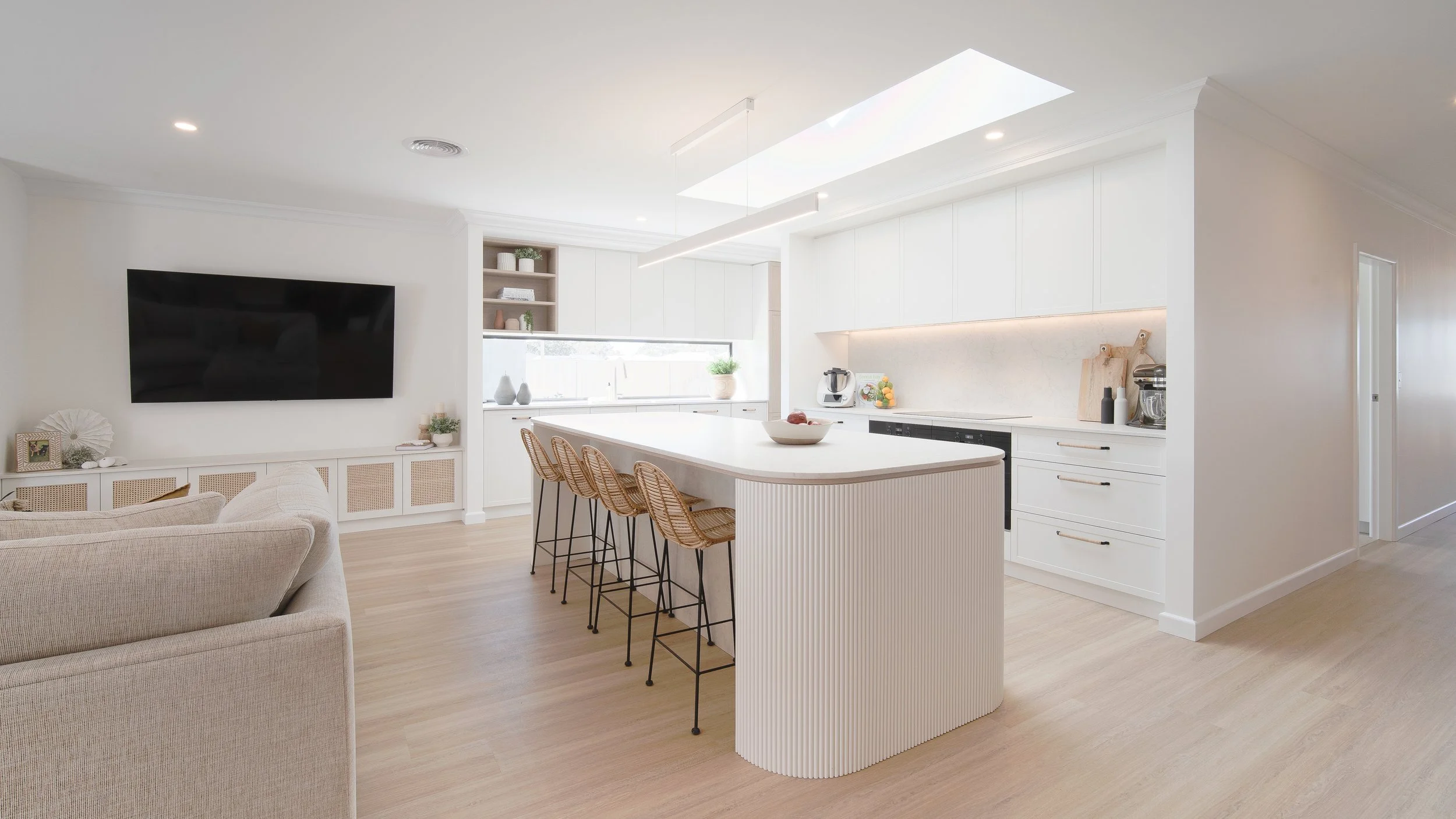 Modern white kitchen with a large island, barstools, and a living area with a wall-mounted TV.