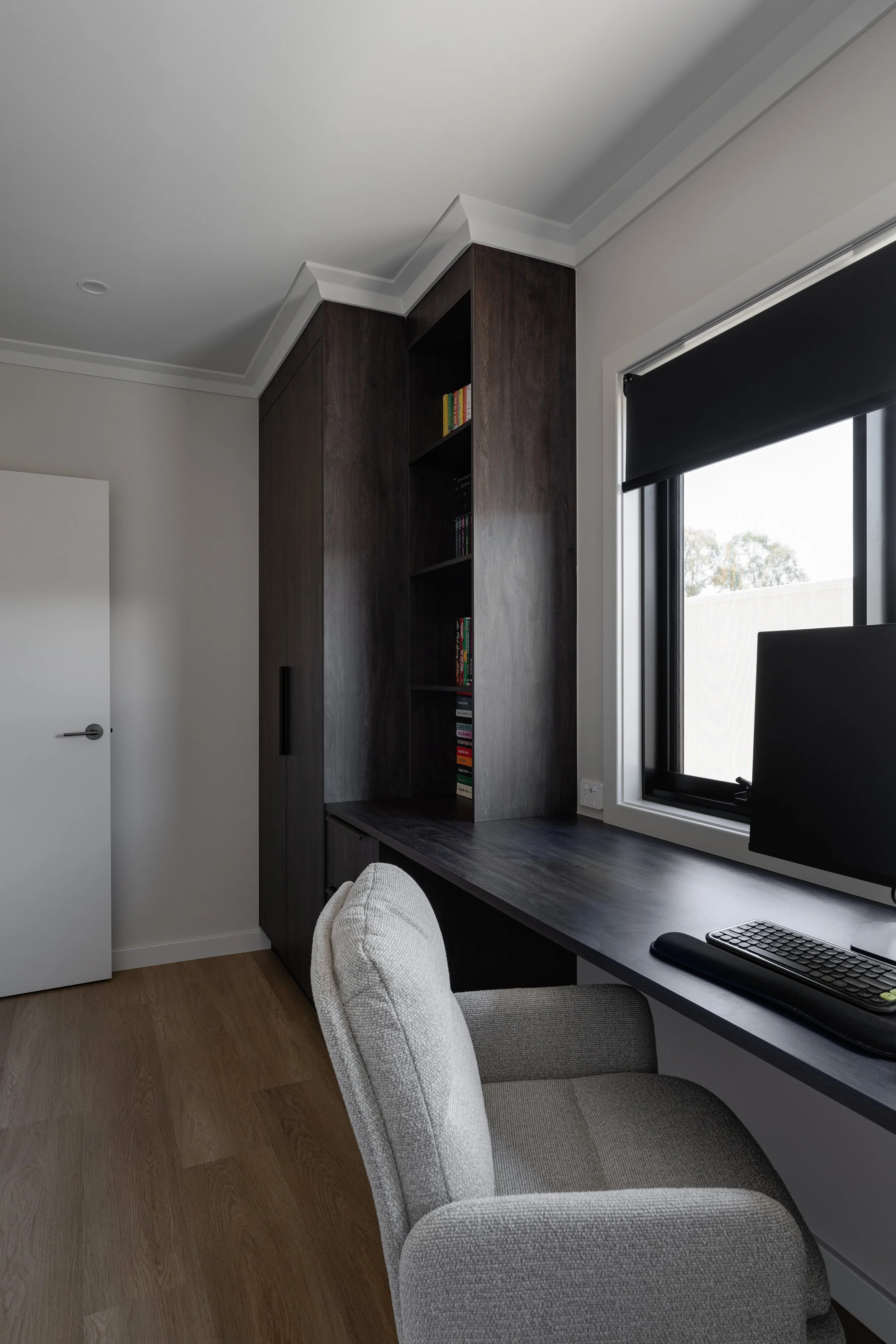 Home office with a built-in dark wood desk and shelving unit, a computer monitor, a keyboard, a mouse, a black window blind, a window, a white door, and a beige armchair.