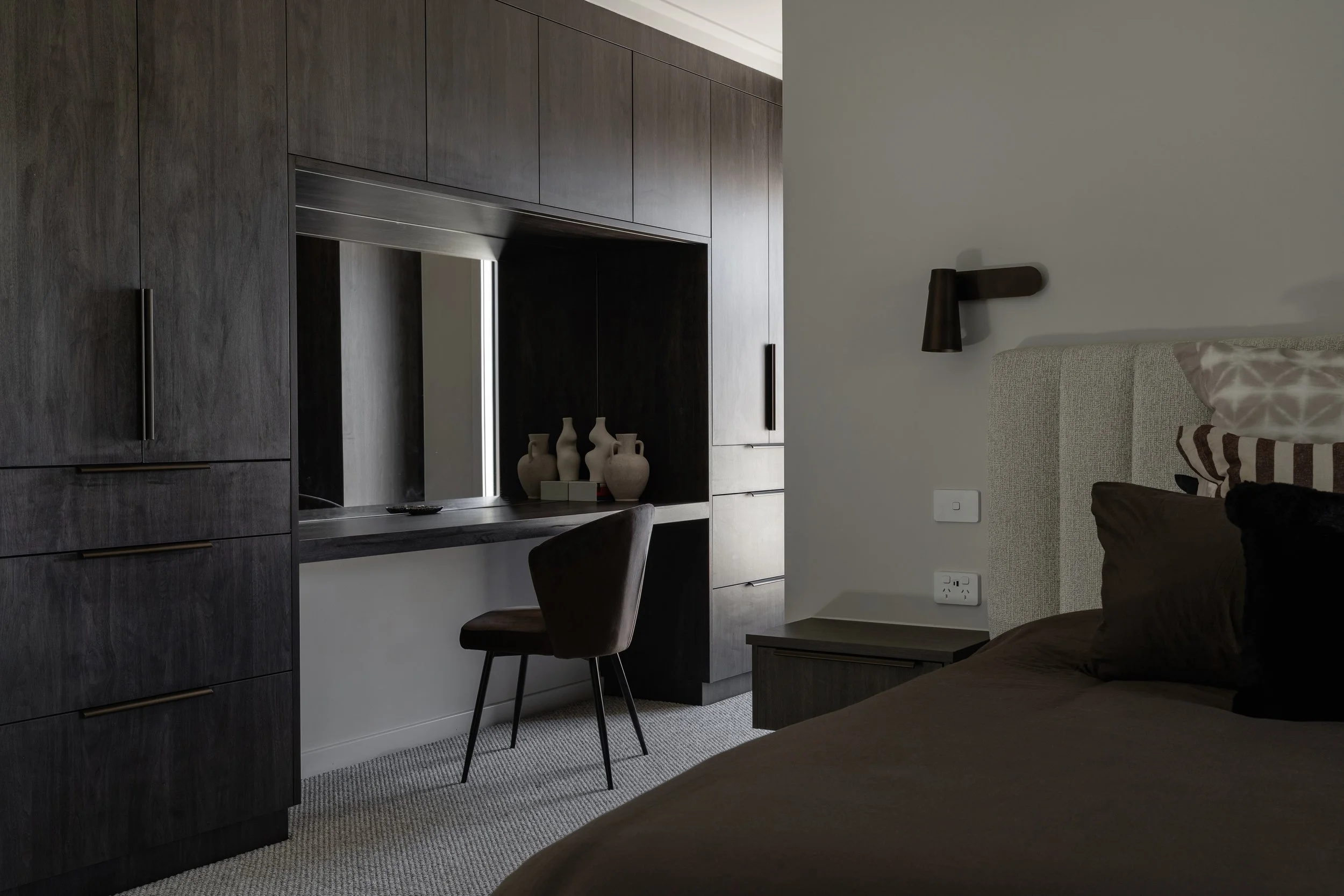 A bedroom with dark wood built-in cabinets, a white wall, a black wall-mounted lamp, a beige upholstered bed with patterned pillows, a table, a chair, and electrical outlets visible.