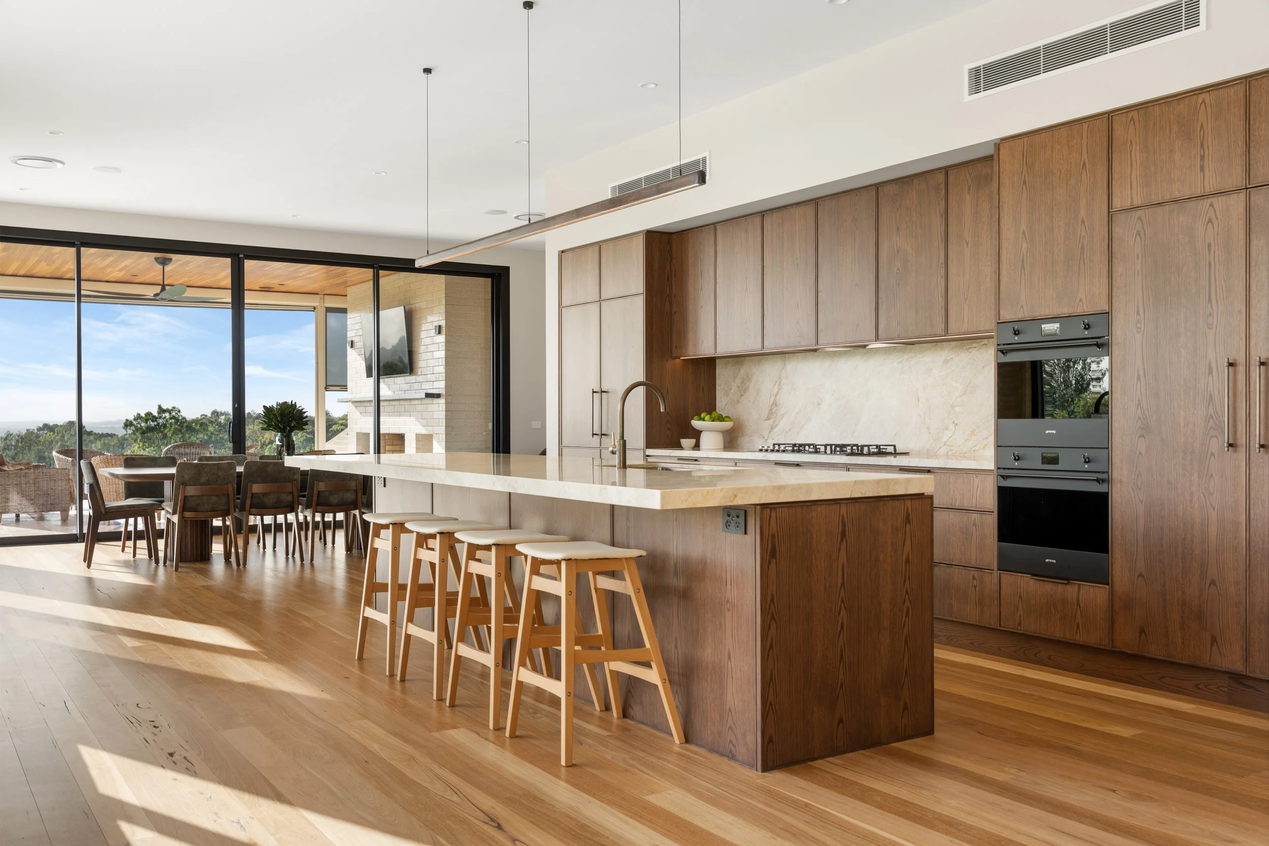 Modern kitchen with wooden cabinetry, a large island with seating, and a view of an outdoor patio with a dining table and chairs through glass sliding doors.