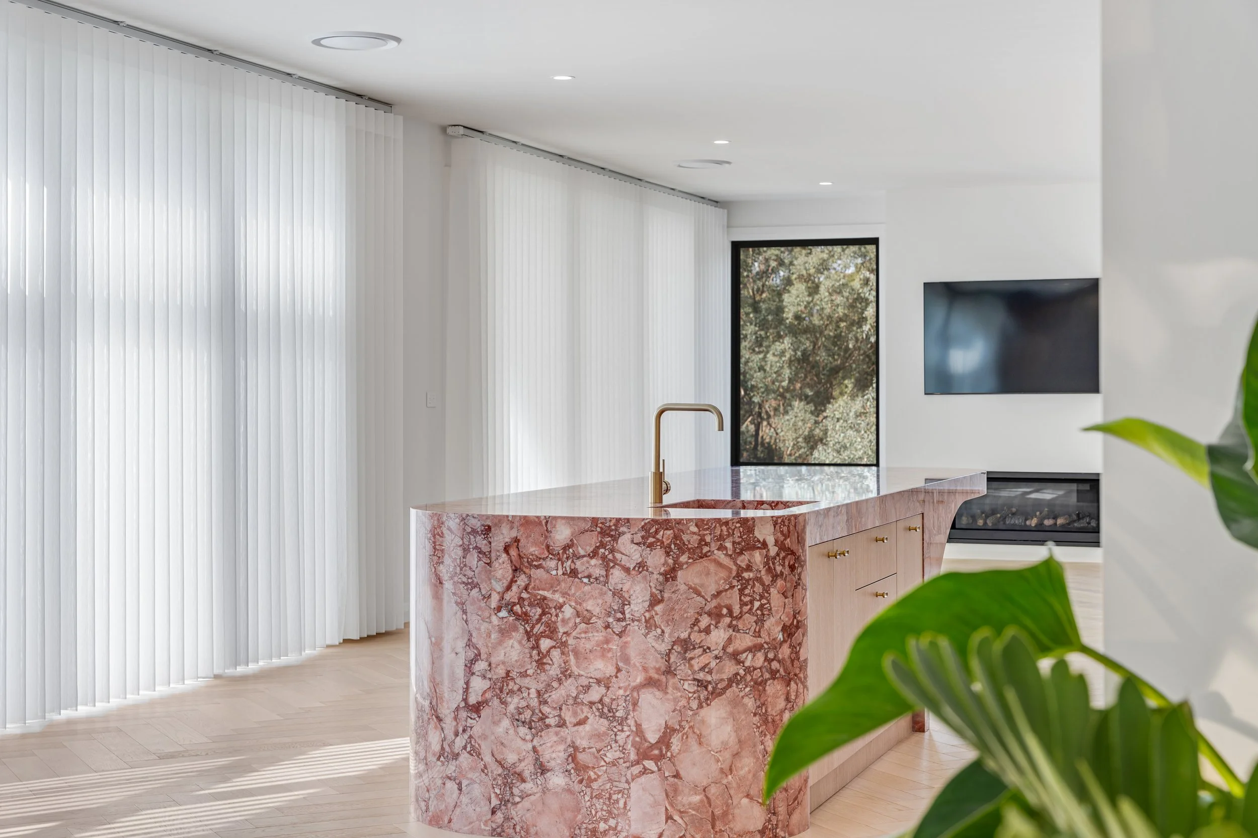 Modern kitchen with a pink marble island, white walls, large window with plants outside, and a black wall-mounted TV.