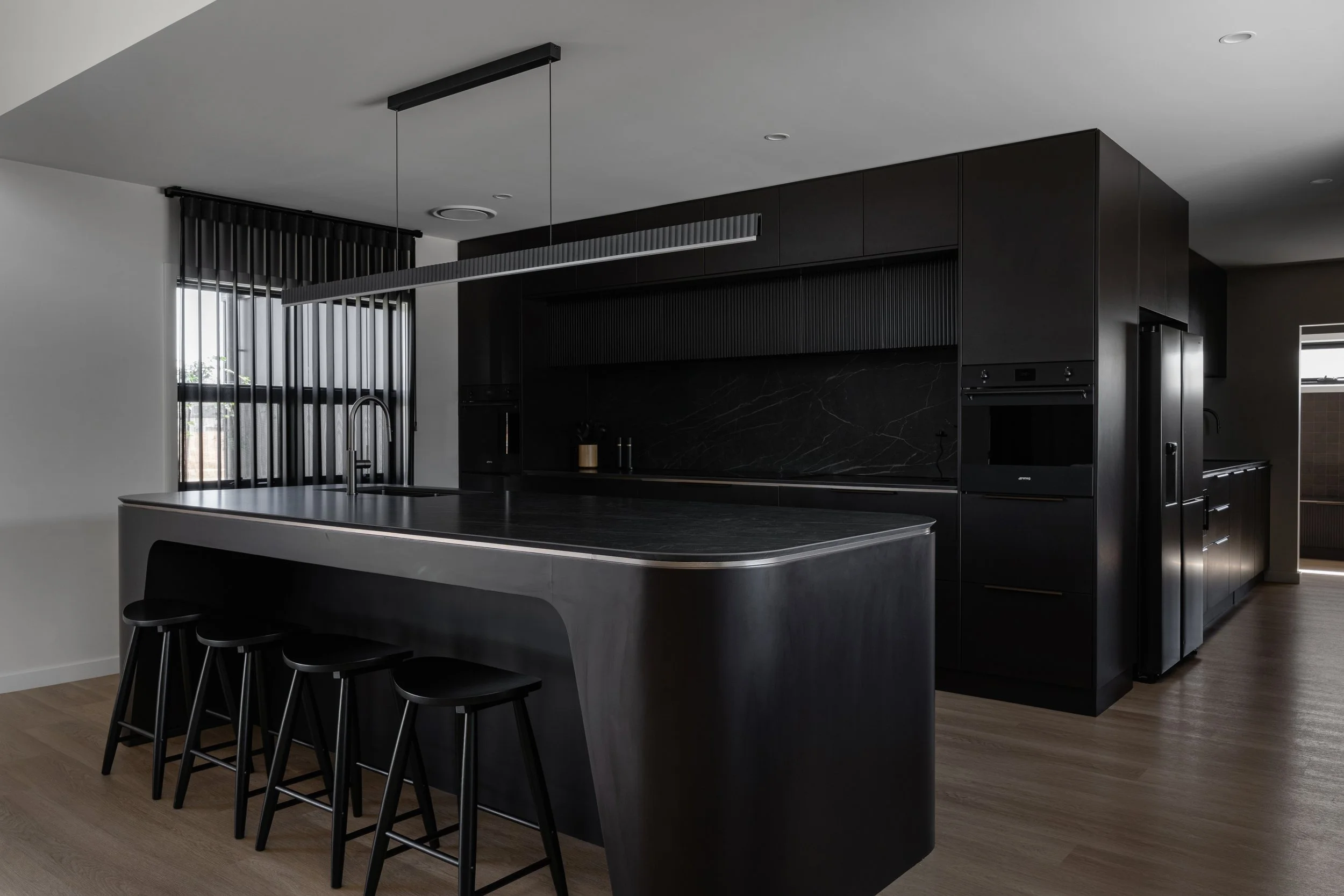 Modern black kitchen with a large island, black cabinets, a black oven, and black bar stools, with natural light coming through windows.