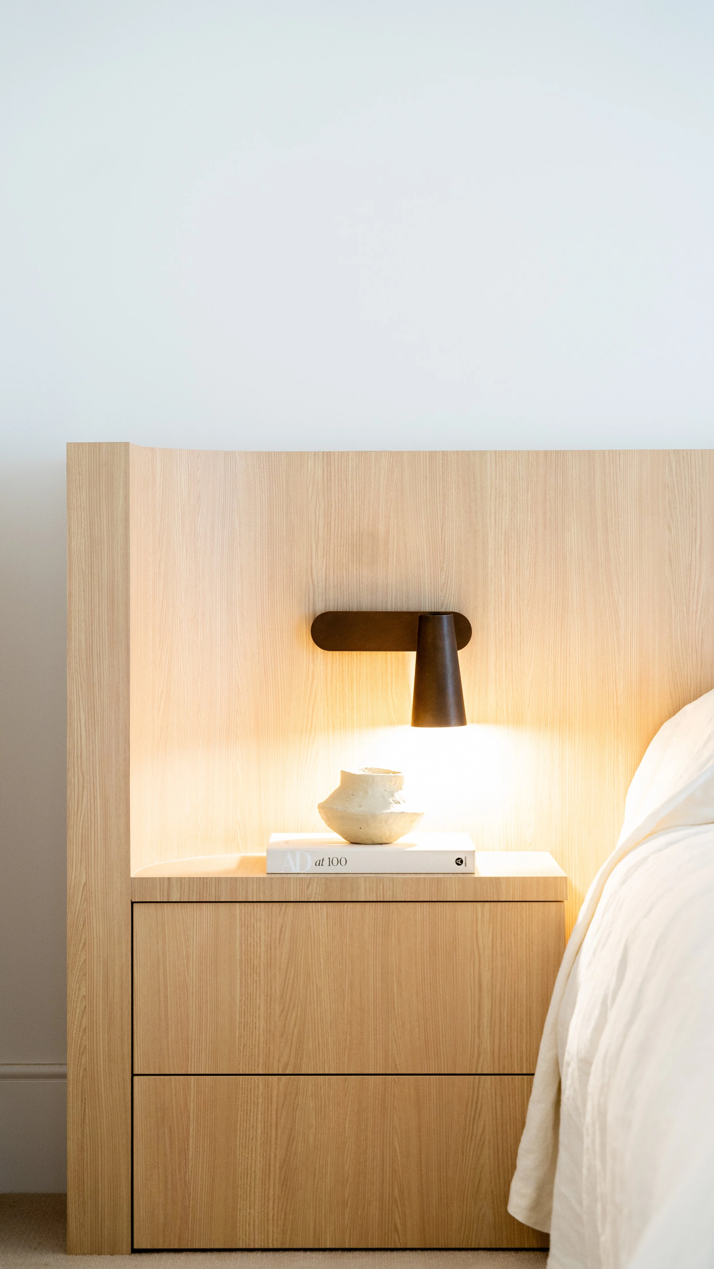A light wood nightstand with a black wall-mounted sconce above it, a white decorative vase, and a white book titled 'AD at 100'. Part of a bed with white bedding is to the right.