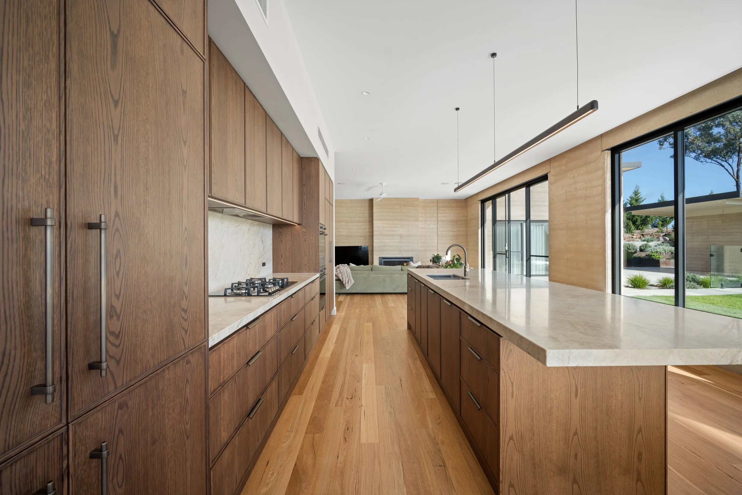 Modern kitchen with wooden cabinets, white countertops, large window with view of outdoor greenery, and open living space in background with sofa and television.