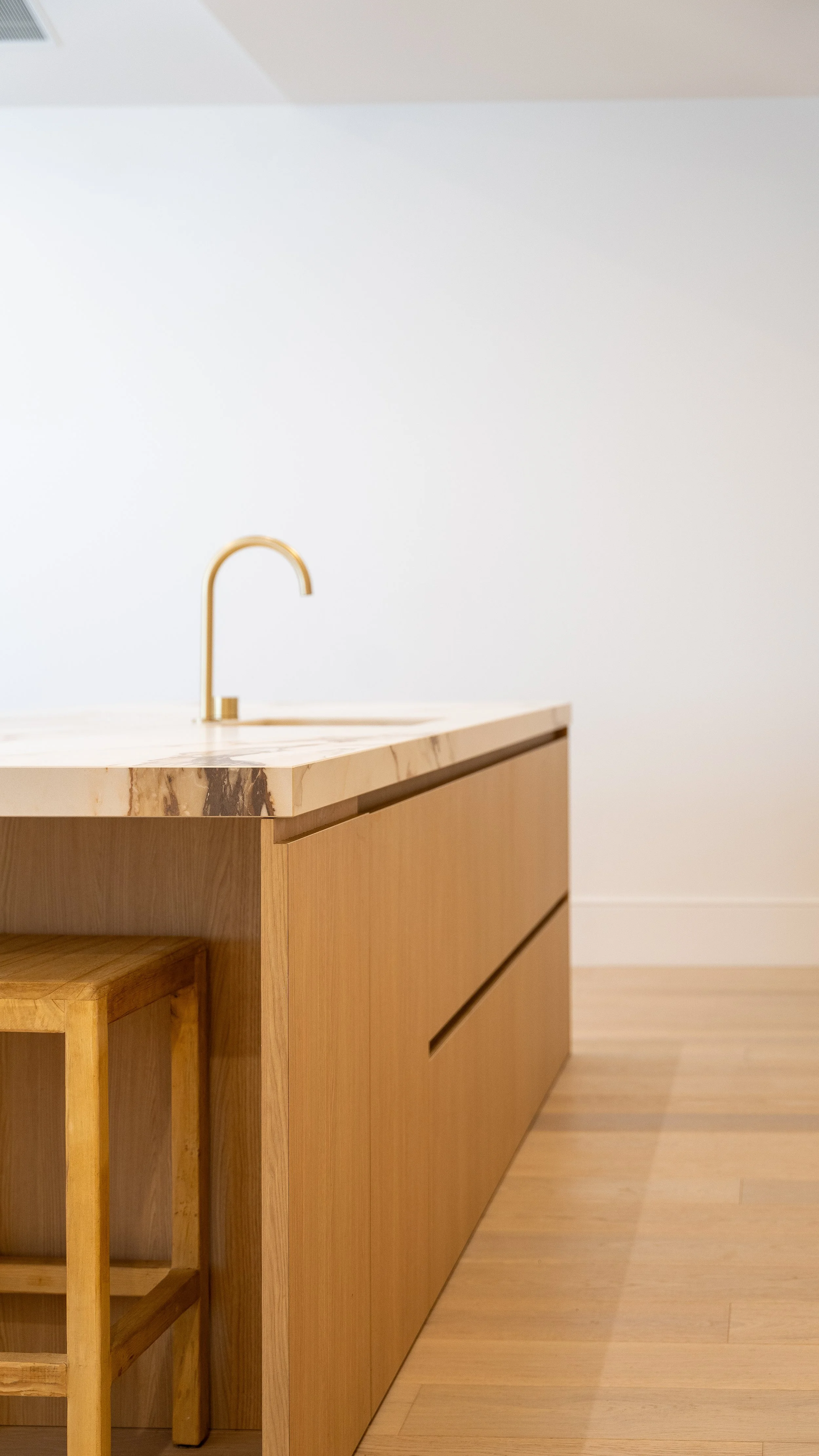 Close-up of a minimalist wooden kitchen island with a marble top, a modern gold faucet, and a small wooden stool beneath, against a plain white wall.