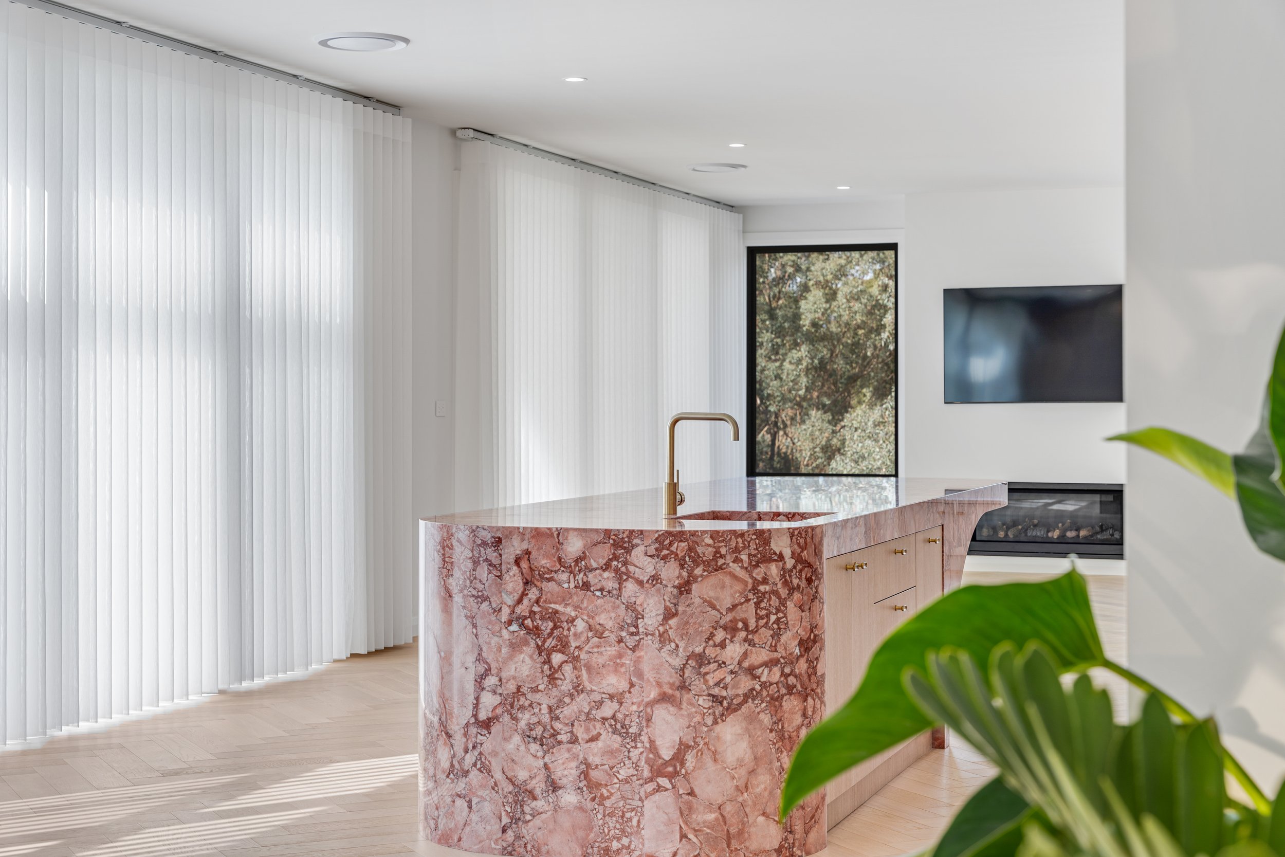 Modern interior kitchen with pink marble island, white vertical blinds, large window, wall-mounted TV, and a green plant in the foreground.