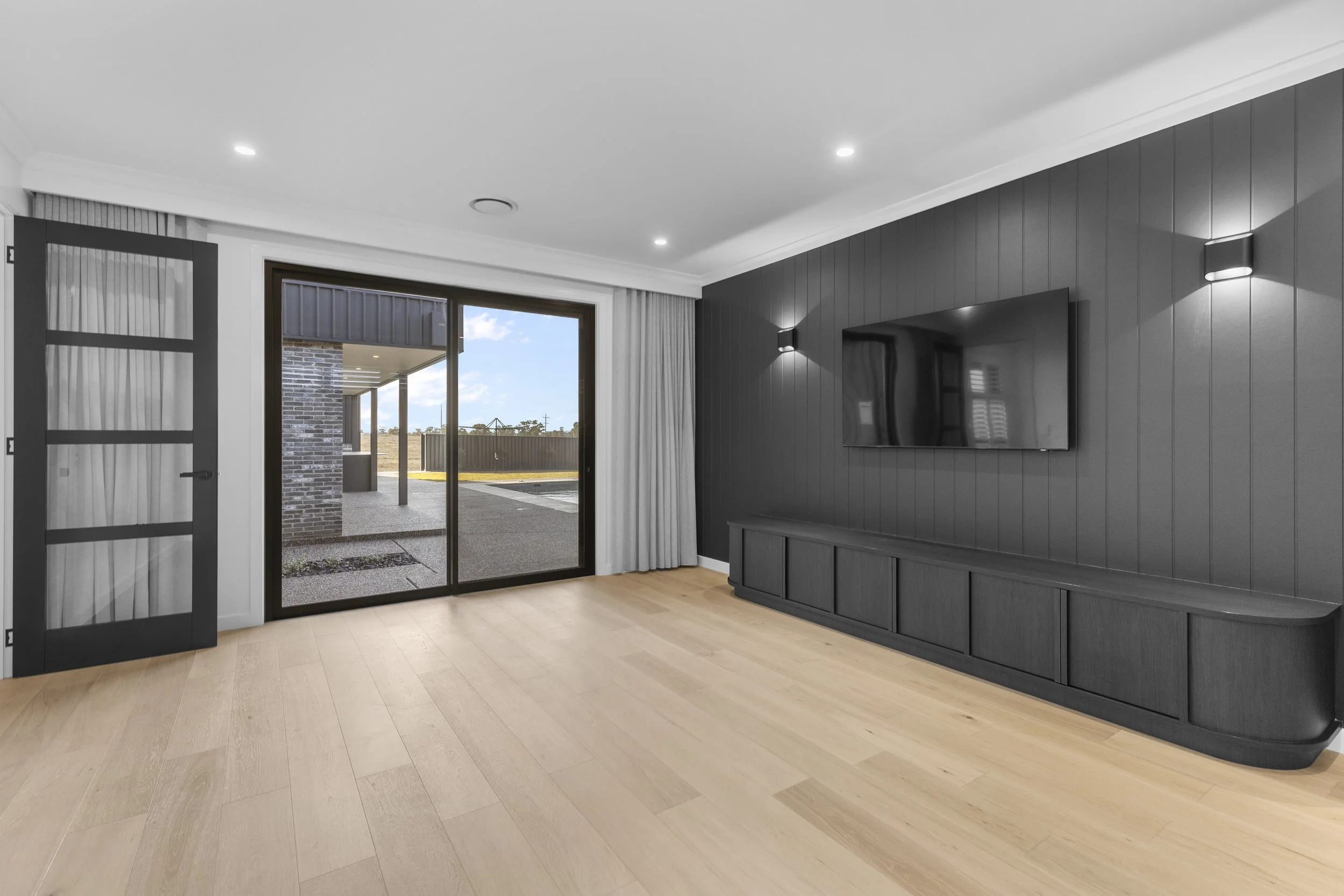 A modern living room with a sliding glass door leading to an outdoor patio. The room has light wood flooring, a dark gray accent wall with a mounted flat-screen TV, and wall sconces. A partially open door with glass panels reveals the outside.