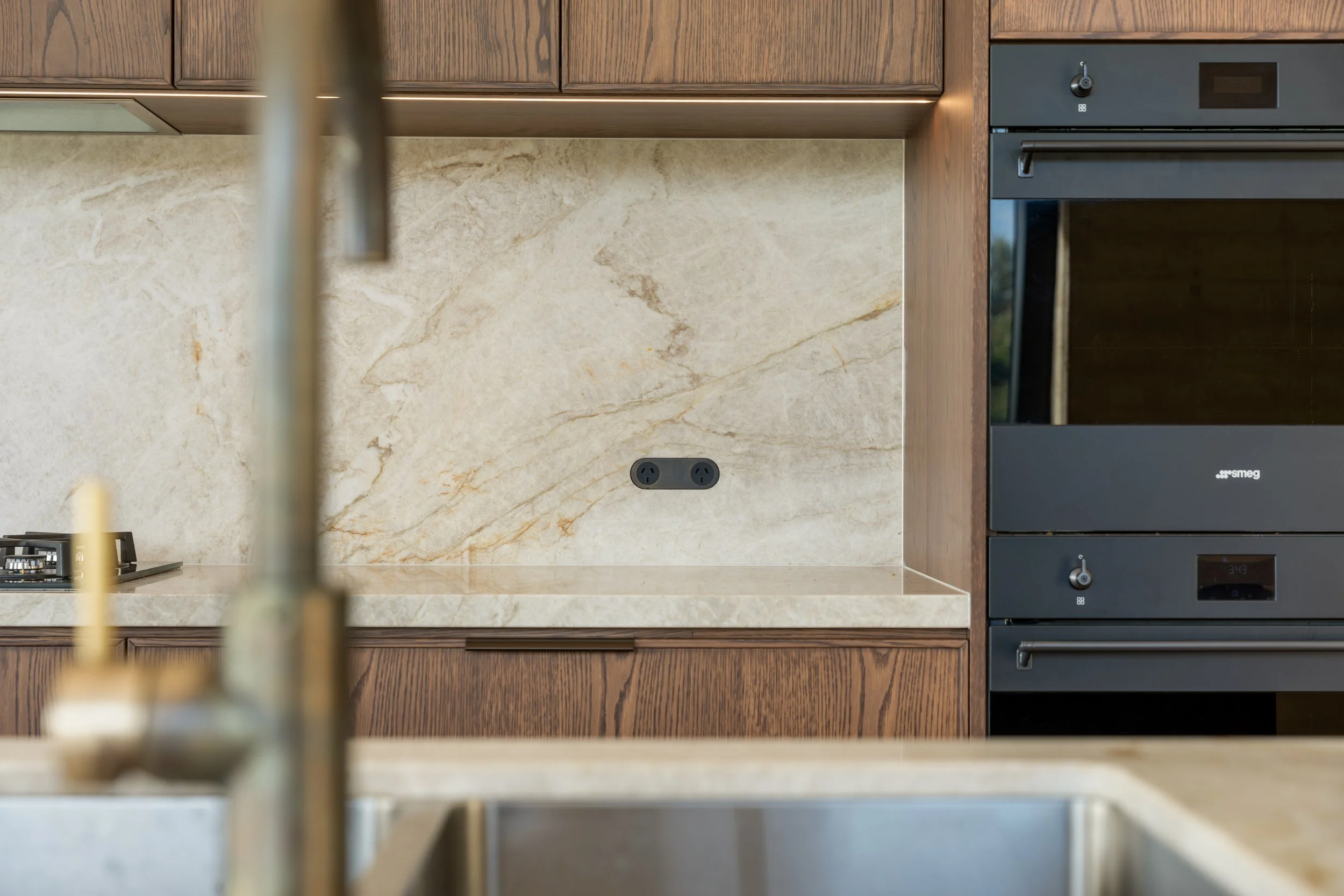 A modern kitchen with wooden cabinets, a beige marble backsplash, and black appliances, including an oven and a stove with a kettle, viewed from behind a sink.