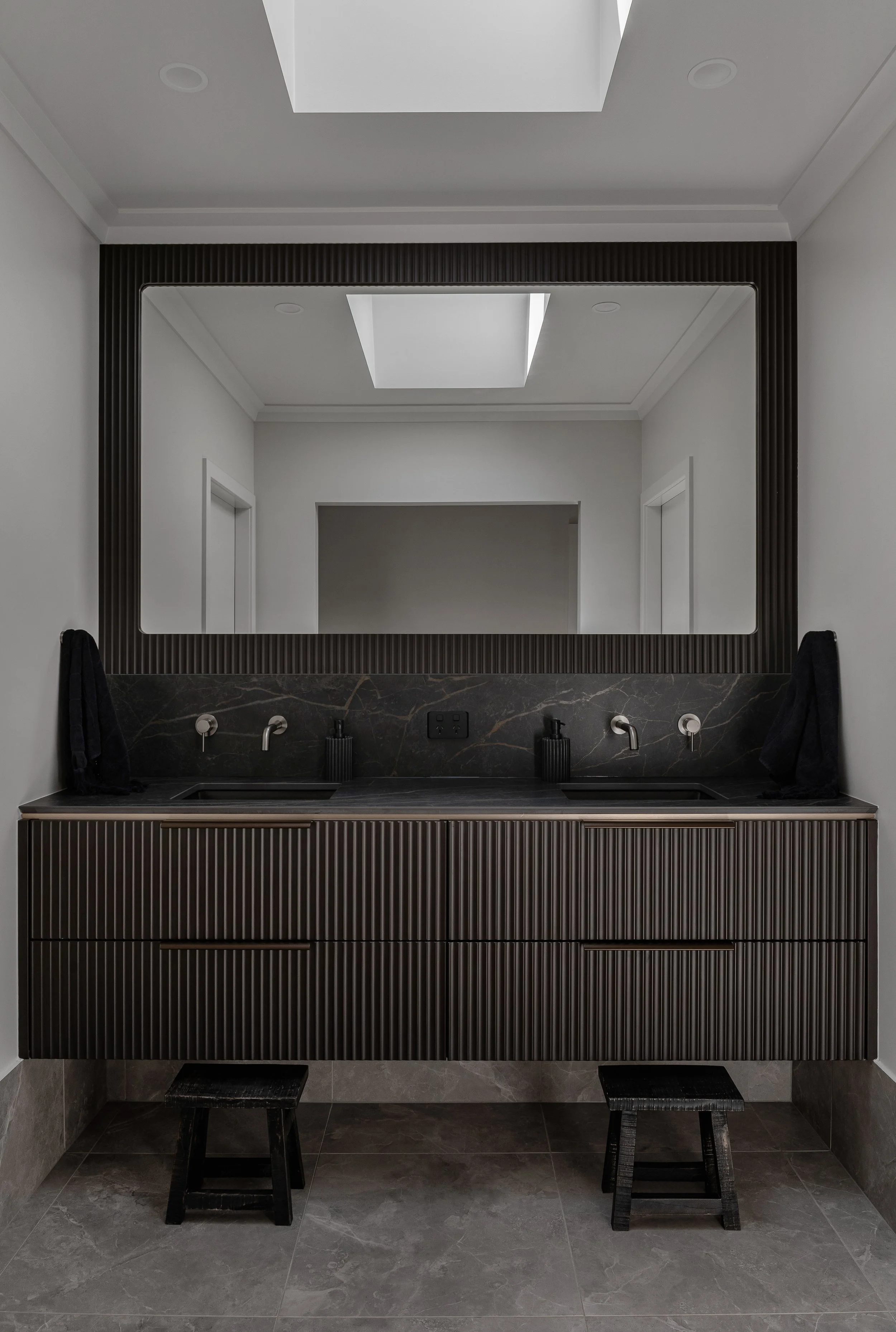 Modern symmetrical bathroom vanity with two sinks, black marble countertop, and textured dark cabinets beneath. Large rectangular mirror above with a black frame, and skylight in the ceiling. Two stools in front of the vanity and two black towels on 