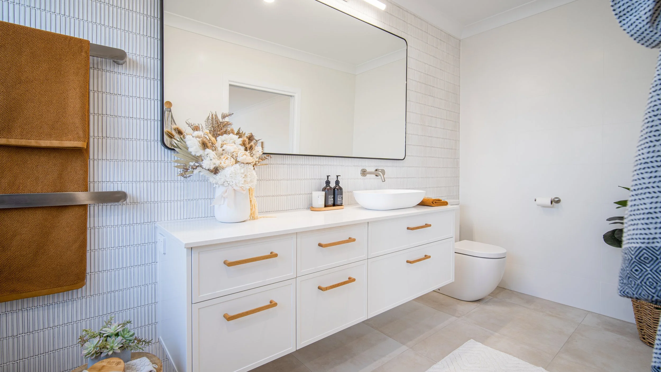 Modern bathroom with white vanity, large mirror, dried floral arrangement, soap dispensers, towel rack, toilet, and a woven basket with a plant.