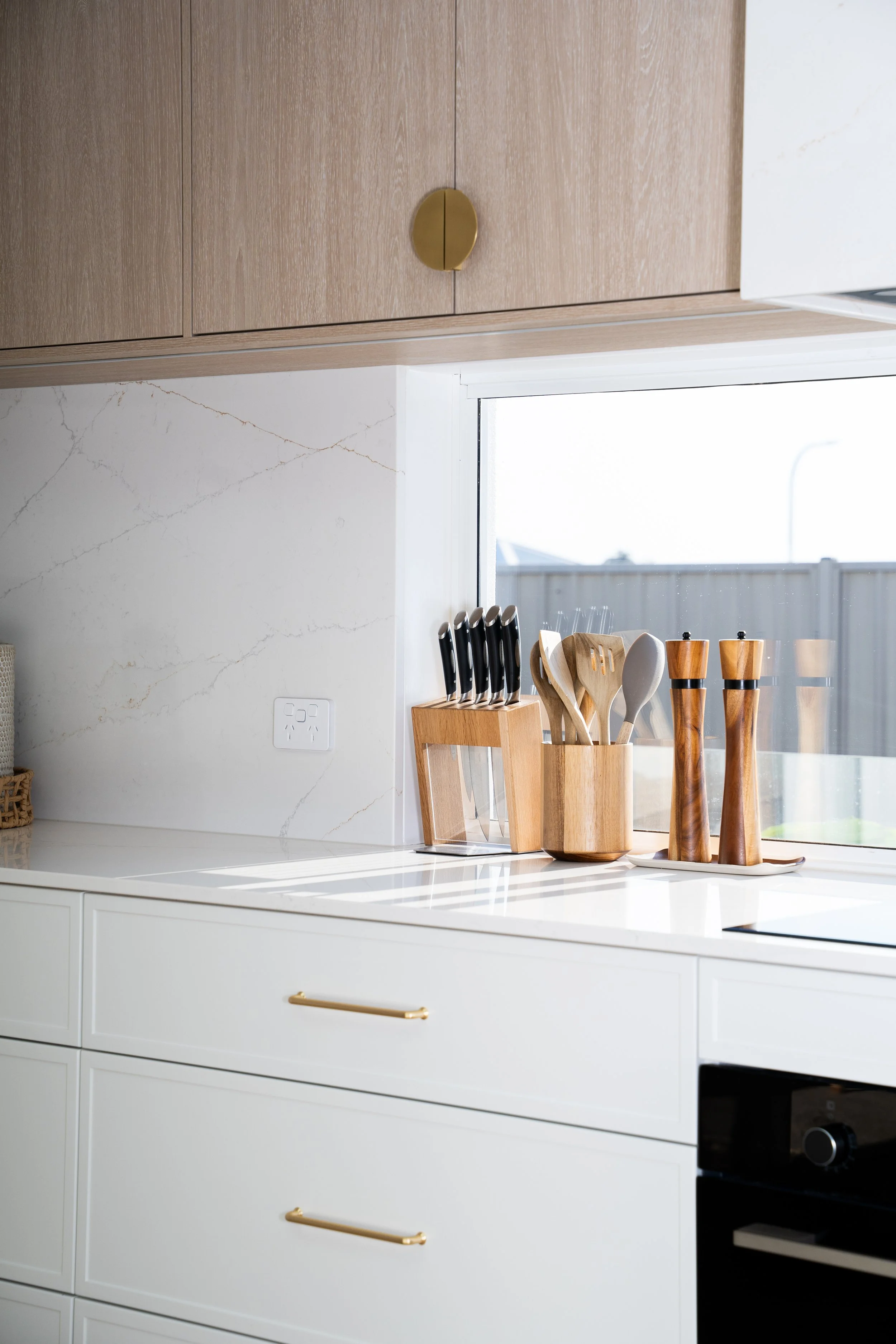Kitchen counter with a white marble surface and white cabinetry with gold handles. On the counter, there is a wooden knife block with multiple knives, a set of wooden cooking utensils, and wooden salt and pepper shakers. A window above the counter sh