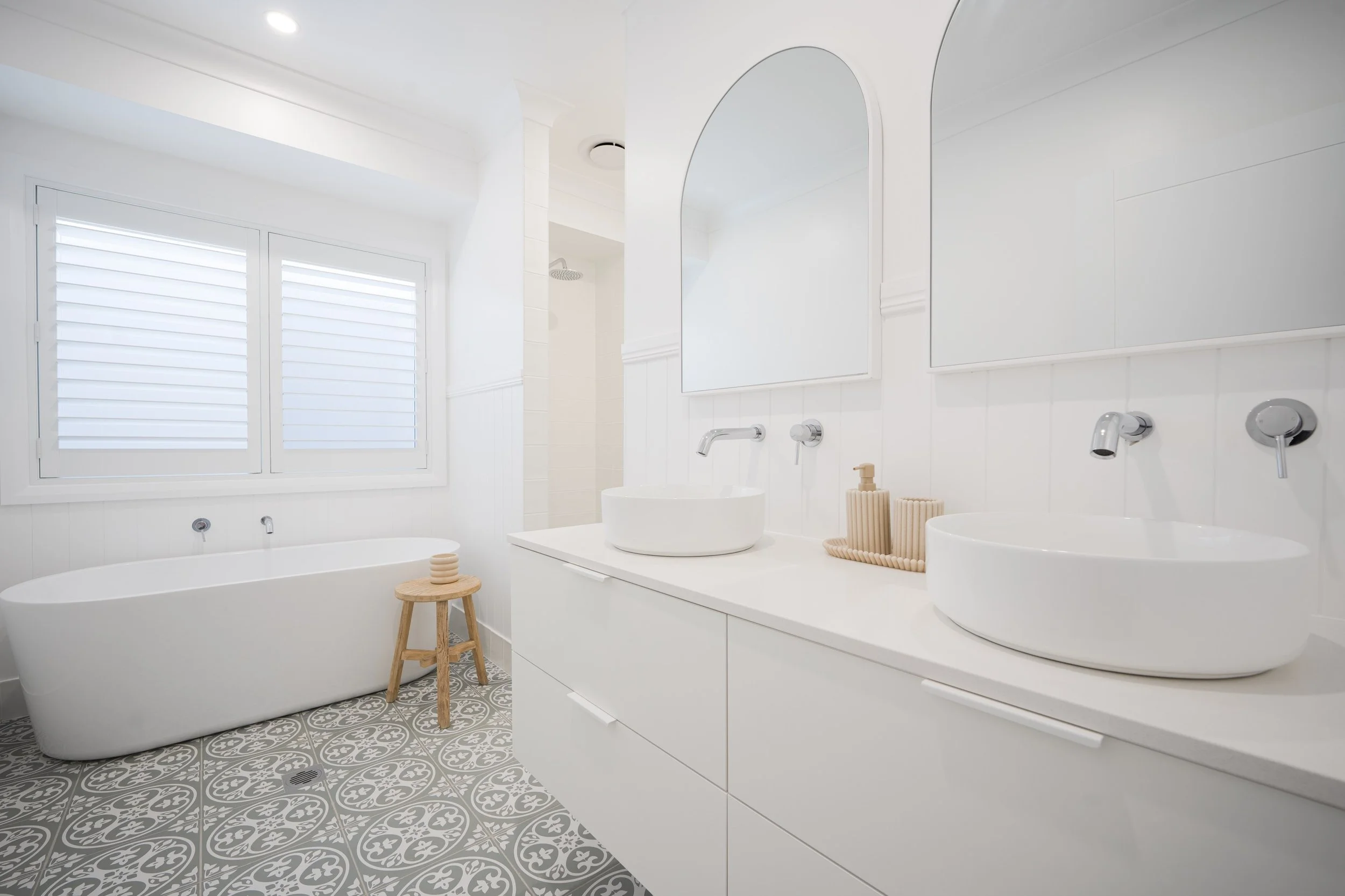 A modern, minimalist bathroom featuring a white bathtub, two white vessel sinks, a large mirror, a window with shutters, patterned tile flooring, a small wooden stool, and neutral decor.