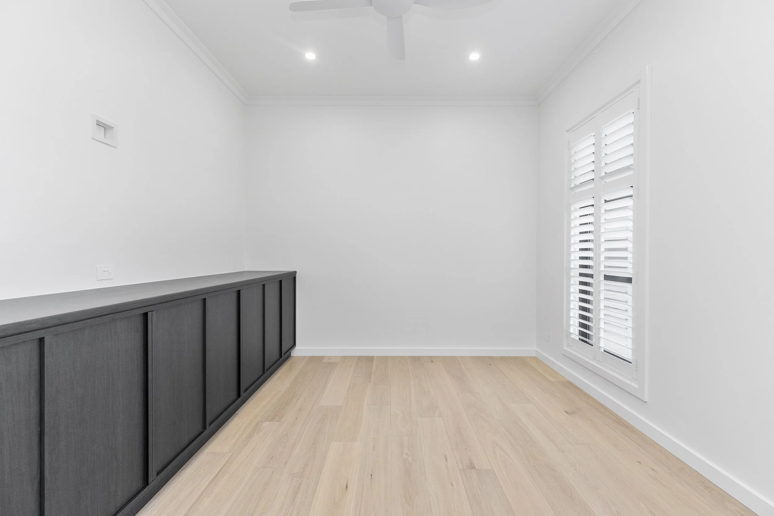 A minimalistic room with white walls, a ceiling fan, recessed lighting, a window with white shutters, and a gray cabinet along one wall.