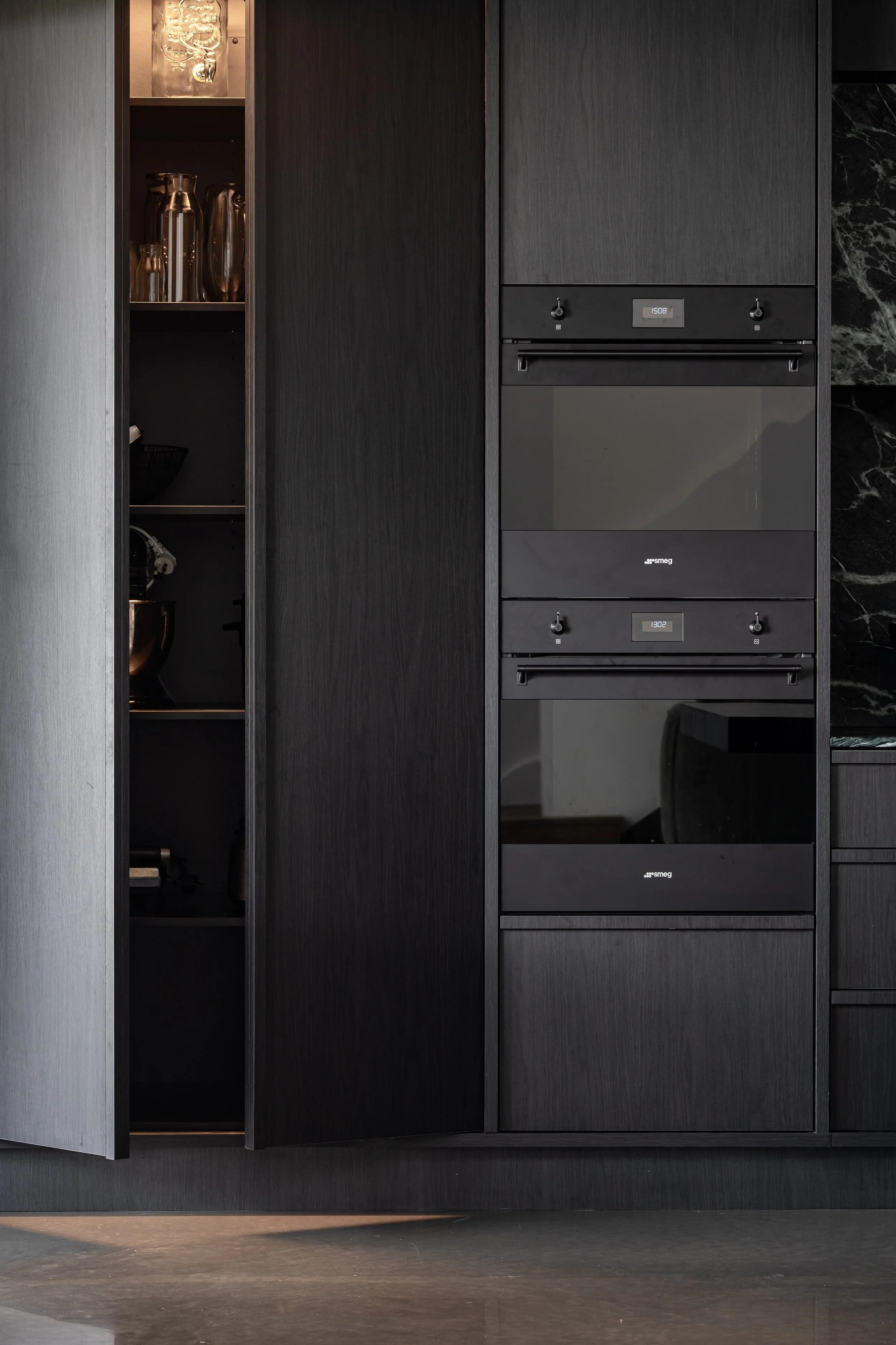 Modern kitchen with black cabinetry and built-in appliances, including an oven and microwave, next to open shelves with metallic kitchenware.