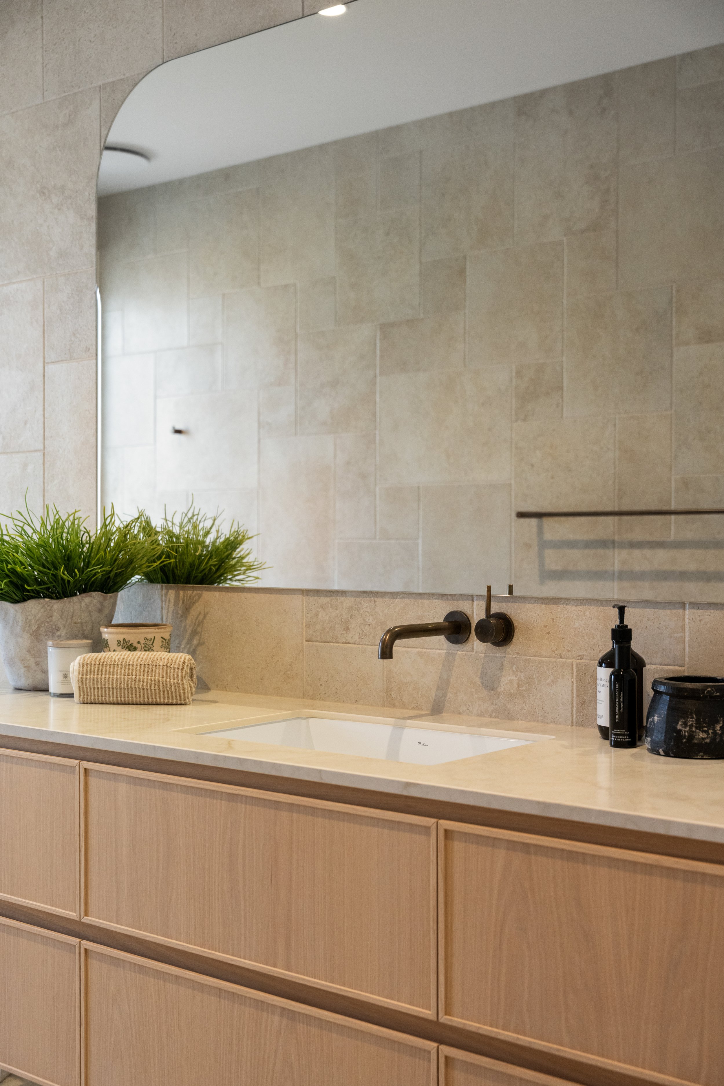 A bathroom vanity with a beige countertop, a white sink, a wall-mounted faucet, a black soap dispenser, a potted plant, and a mirror reflecting beige tiled wall.